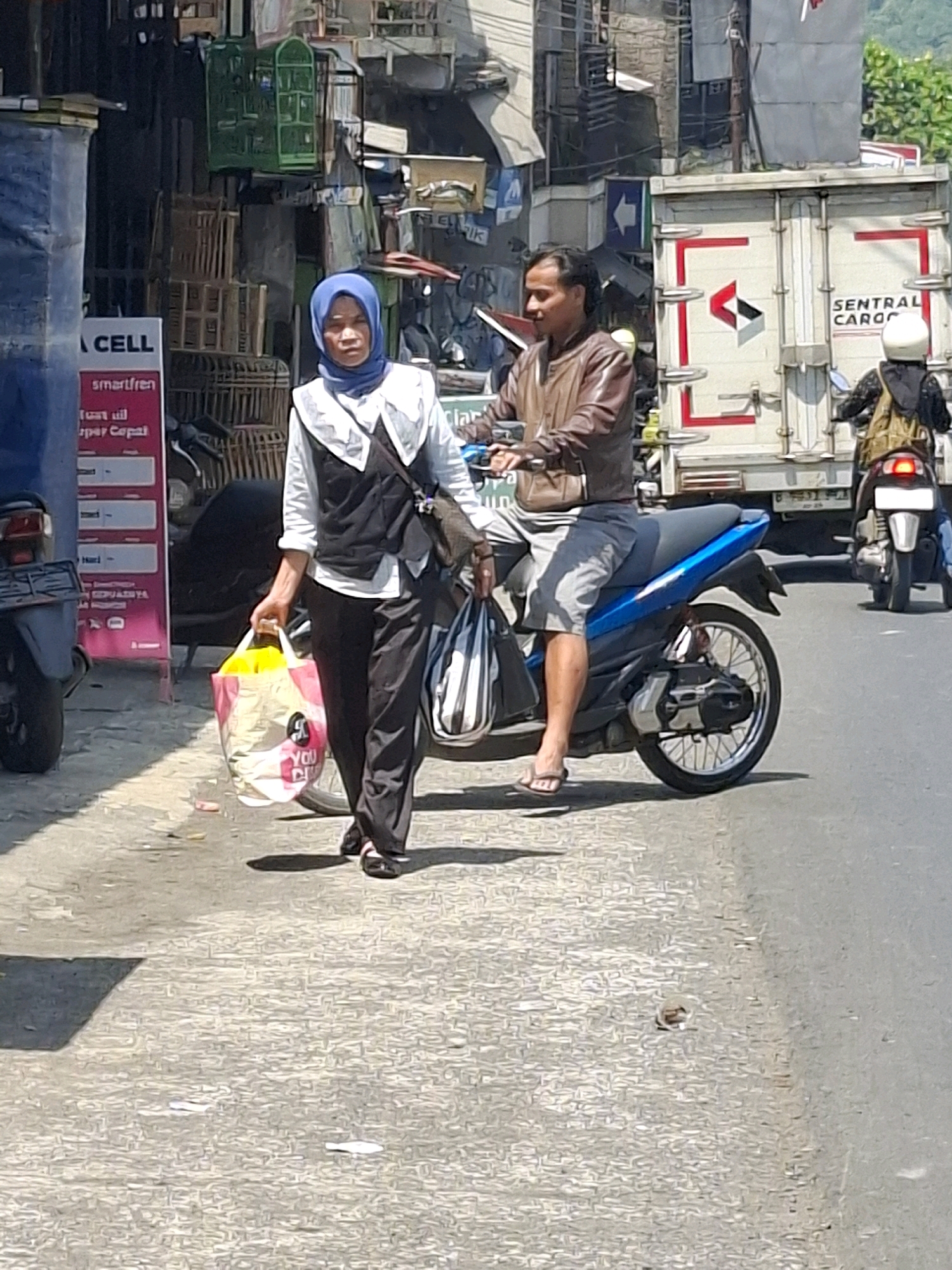 In Cikahuripan, Indonesia, a woman wearing a blue hijab and a white and black vest walks on a paved street, carrying two shopping bags. One is a large pink and yellow bag with "YOU CAN BE SMART" printed on it, and the other is a striped black and white bag. She is walking next to a blue motorcycle on which a man in a brown leather jacket and grey shorts is seated, looking to his left. Behind them, a white truck with "SENTRAL CARGO" written on its side is parked. The environment appears to be an urban area with buildings and stalls lining the street. The image is taken during daylight hours.