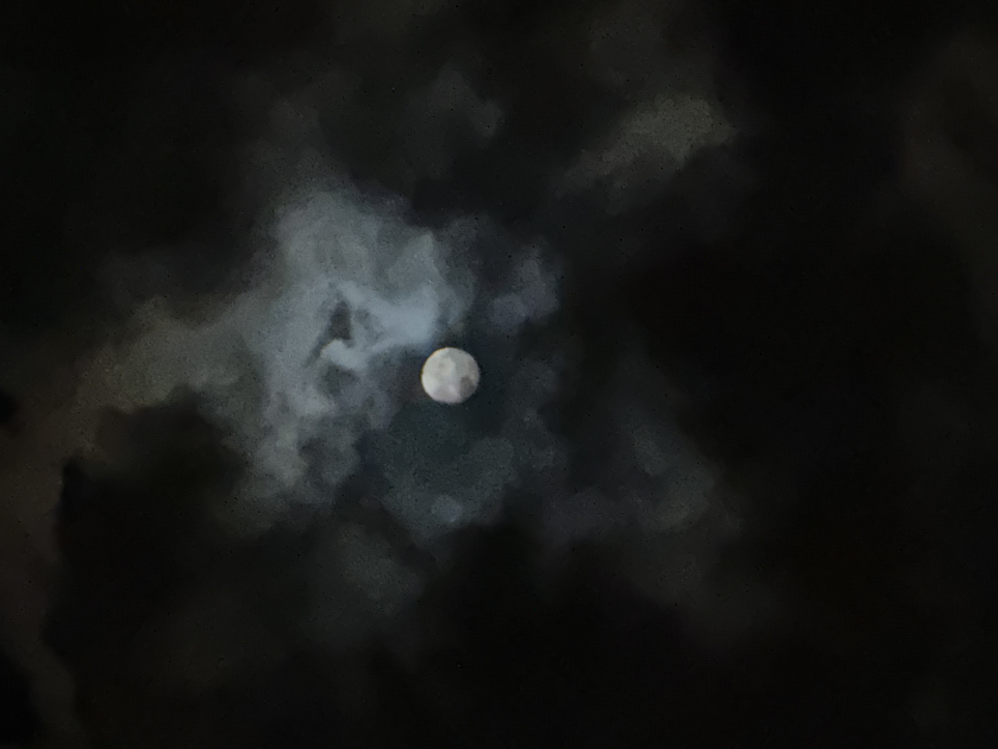The image depicts a full moon seen through clouds at night. The moon is slightly off-center, positioned towards the middle right of the frame. The clouds are dense and dark, obscuring the moon's light and details. There is no information about the location being Yerevan, Armenia, so I will exclude that. There are no people, subjects, objects, actions, or interactions visible in the image other than the moon and clouds. The environment is dark and cloudy.