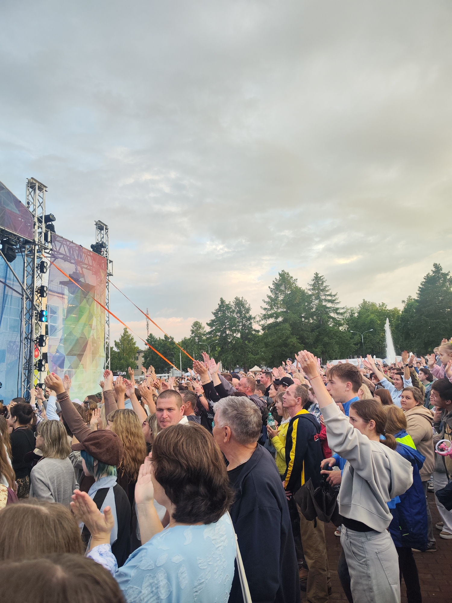 The image depicts a crowd of people with their hands raised, attending an outdoor event in Chaikovsky, Russia. In the background, a stage structure with lighting and a large, colorful banner is visible. Trees and a fountain can also be seen. The sky is overcast. The people in the foreground and mid-ground are diverse in age and gender, dressed in casual clothing. Many are looking towards the stage, suggesting they are watching a performance or presentation. The overall atmosphere appears to be one of engagement and enjoyment.
