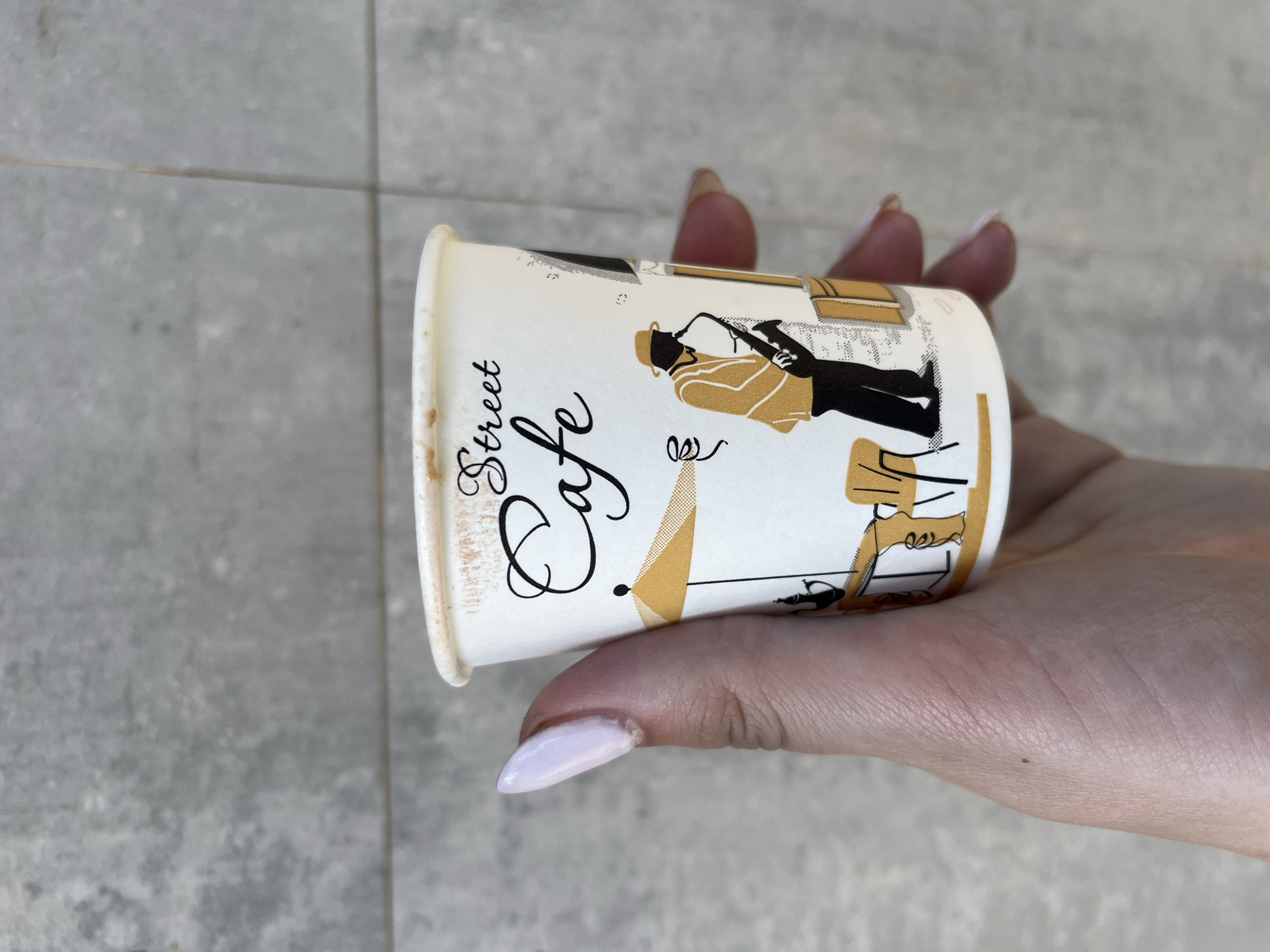 A person is holding a white paper coffee cup in their hand against a light gray tiled background. The cup has a design depicting a street cafe scene in shades of black and gold. The text "Street Cafe" is prominently displayed on the cup. The person's hand is slightly tanned and their fingernails are painted a pale lavender color. The background consists of large square tiles. No location information about Burgas, Bulgaria is discernible.
