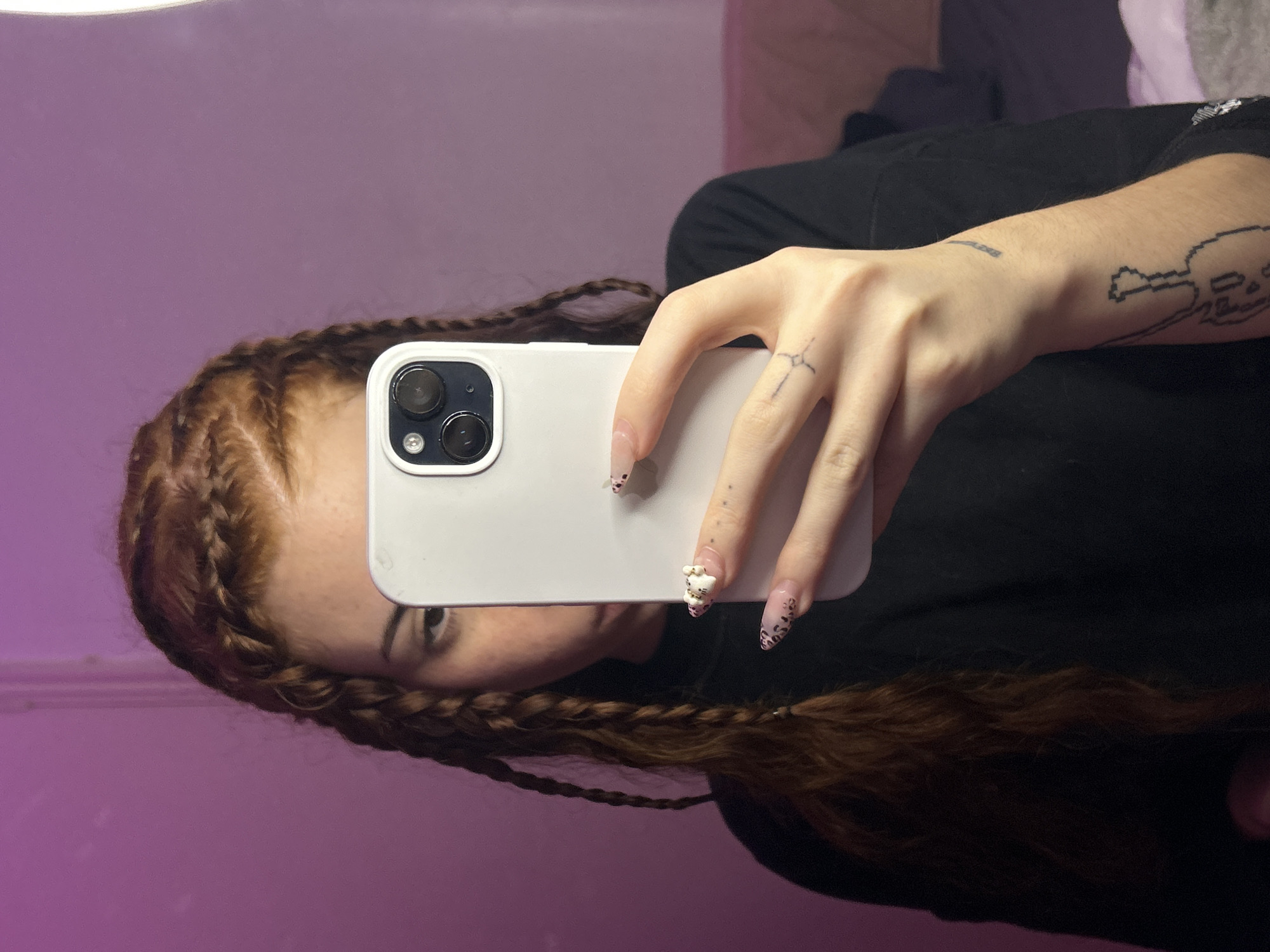 A woman with red-brown braided hair holds a white smartphone, partially obscuring her face. She is wearing a black shirt and has tattoos on her hand and forearm. The background appears to be a light purple wall. The image does not contain enough information to determine the location as Buenos Aires, Argentina.