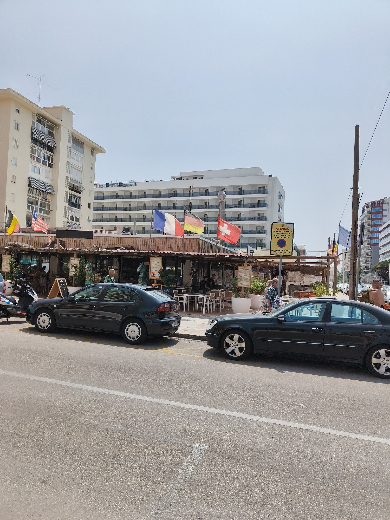 The image captures a street scene in Gandia, Spain, on a sunny day, likely near the beach given the presence of flags from various European countries flying outside a row of establishments. Two dark-colored sedans are parked on the street in the foreground. The left car, a dark hatchback, is parked closer to the establishments. The right car, a dark sedan with a sunroof, is parked further to the right. Behind the cars, there is a promenade or sidewalk area with tables and chairs set up outside open-air establishments, suggesting a commercial strip with restaurants or cafes.
Several flags are visible, including the flags of Belgium, the United States, France, Germany, and Switzerland, adding a cosmopolitan feel to the location. A yellow traffic sign with red lettering is posted on a pole in the middle ground. People are visible in the background, some walking and others seated at tables. The architecture in the background includes multi-story buildings, one of which appears to be a hotel with balconies. The sky is clear and bright, indicating fair weather. A wooden utility pole stands on the right side of the image.