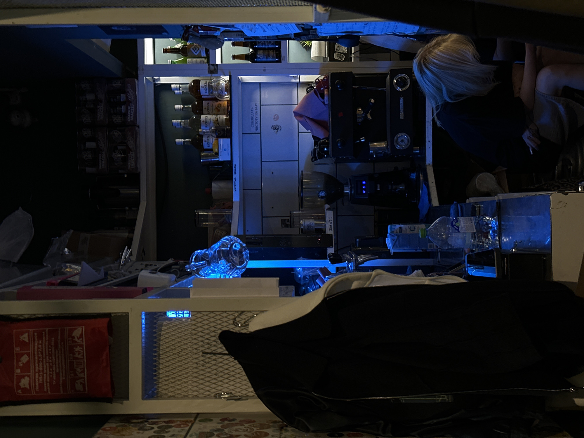 Here is a forensic description of the image.
The scene is dimly lit and appears to be the interior of a bar or cafe, potentially after hours. A fair-haired person is partially visible, seated near an espresso machine. Shelves are stacked with bottles, jars, and boxes, likely liquor and beverage supplies. The environment appears cluttered with loose items strewn about, including water bottles and a red bag. A black jacket lies in the foreground. The overall impression is one of disarray and after-work activity. The location is Unknown, Unknown.
