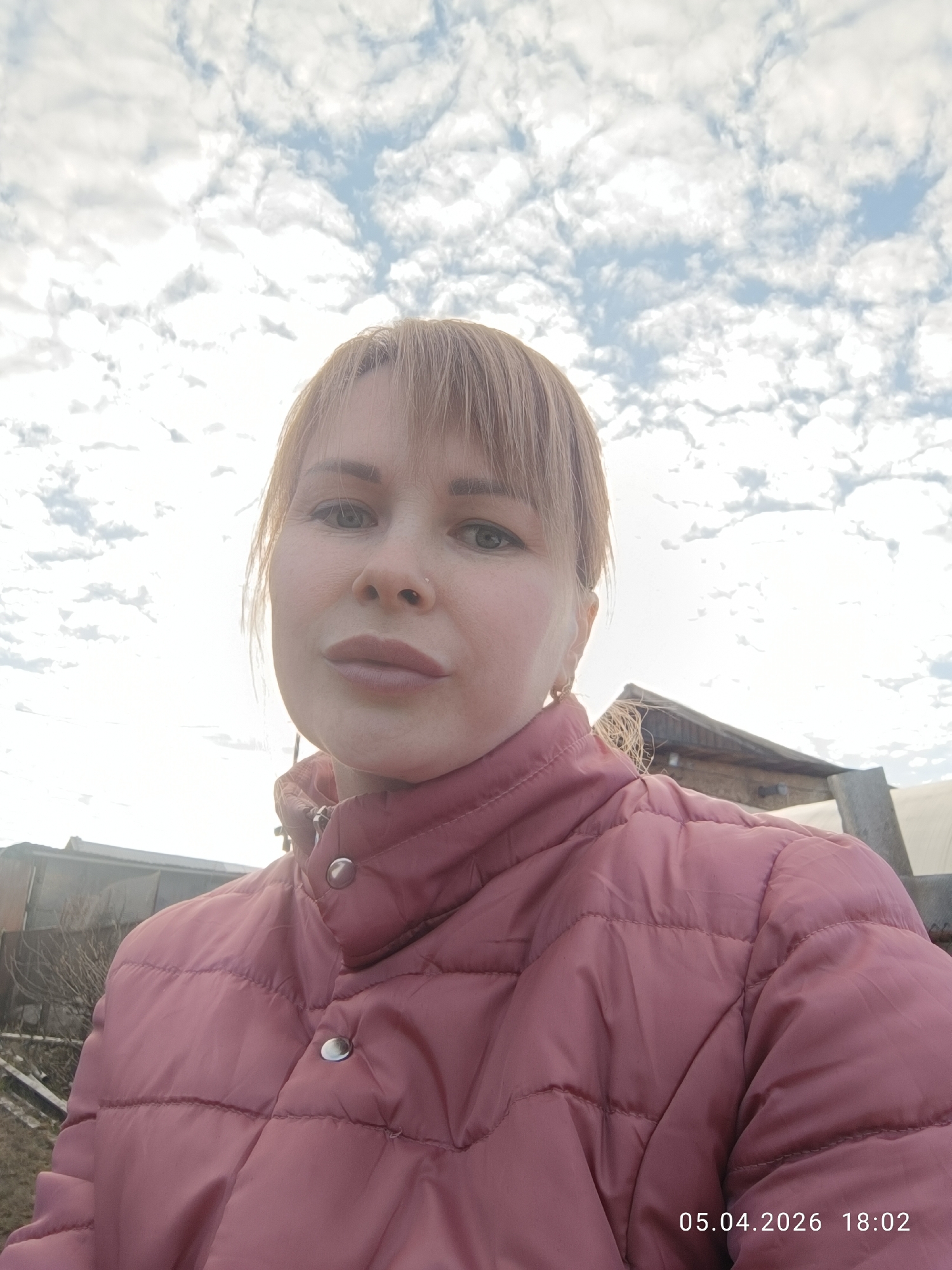 A selfie photograph taken by a woman, showing her from the chest up, with the sky visible overhead. She is wearing a pink quilted jacket. A building and some vegetation are visible in the background. The image includes a timestamp: 05.04.2026 18:02. The location is stated as Chaikovsky, Russia.