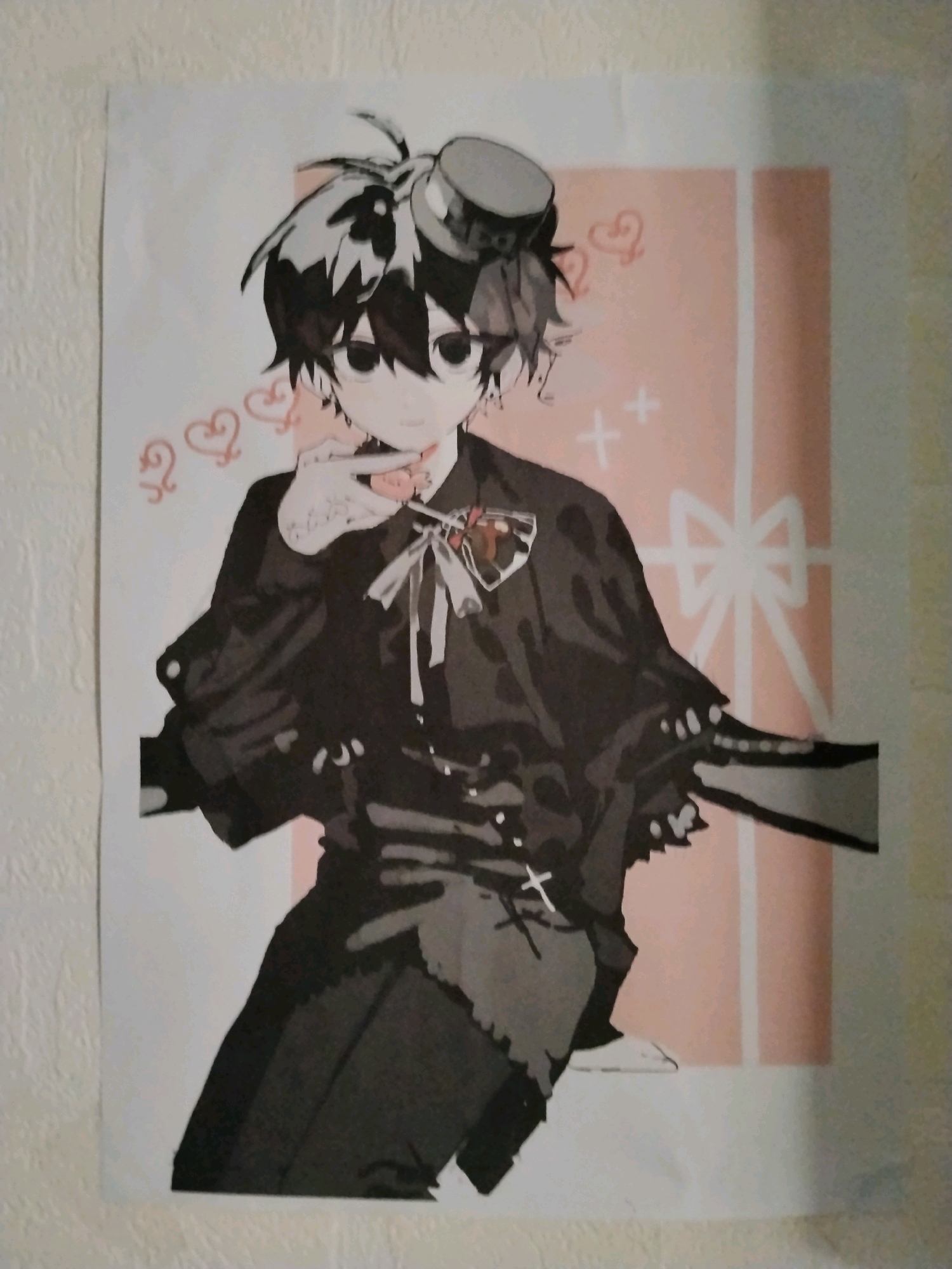 A poster of an anime character, with dark, spiky hair, a top hat, and a black outfit with a red bow, is displayed on a textured wall in what appears to be a residential setting. The character is depicted in a stylized, somewhat Gothic manner, with large, dark eyes and a hand raised to their face, perhaps gesturing or holding something. The background of the poster features a soft, peachy color with stylized hearts and crosses, suggesting a thematic element of romance or perhaps a darker, playful aesthetic. The lighting in the image is ambient and diffuse, indicating it was taken indoors with artificial or indirect natural light. The poster itself is slightly crumpled in places, suggesting it may have been recently hung or is not perfectly preserved. There are no identifiable landmarks or elements that would specifically place this scene within Yogyakarta, Indonesia; the description is limited to the visual content of the provided image and its crops.