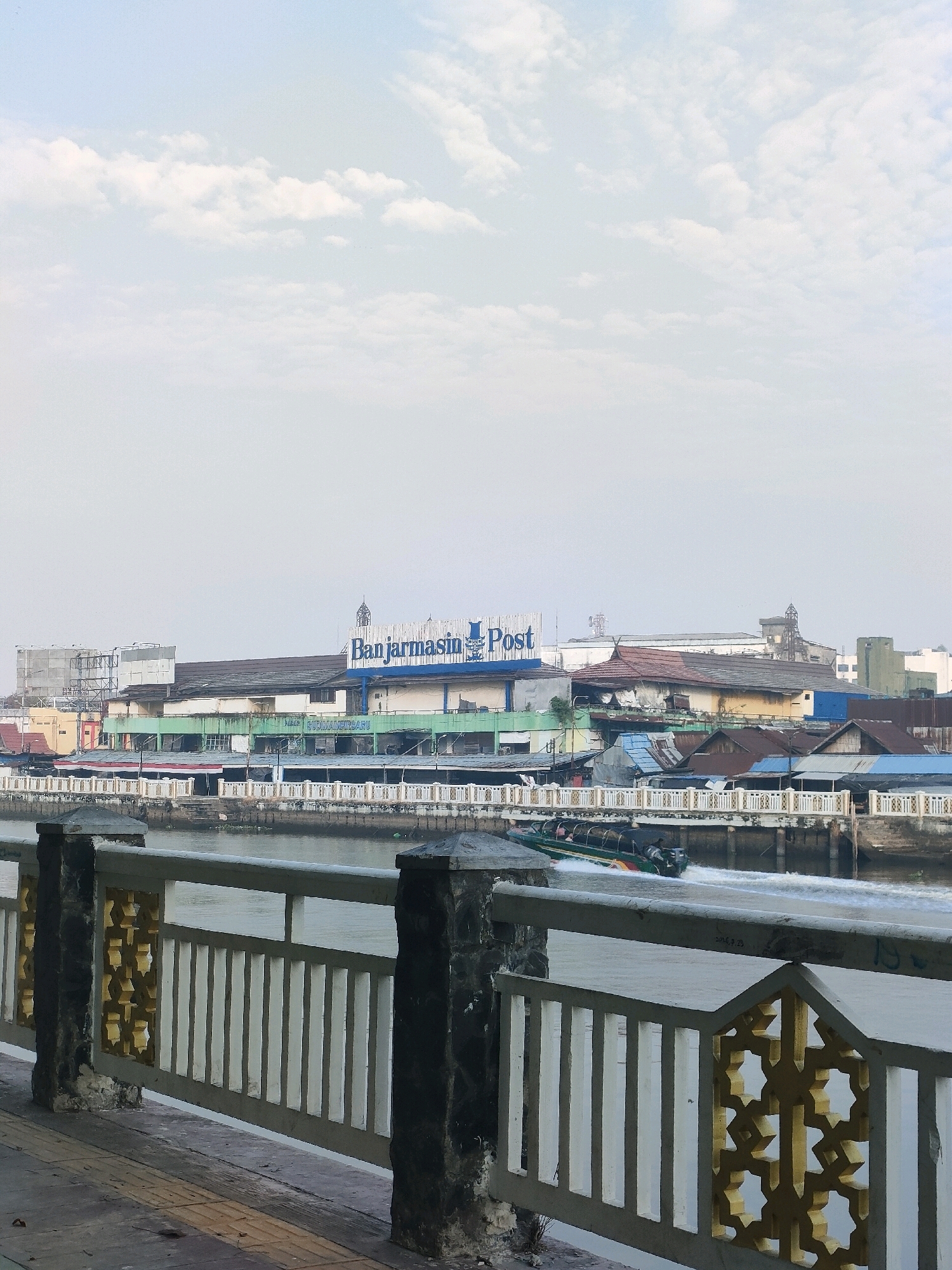 This image depicts a waterfront scene in Banjarmasin, Indonesia, likely along the Martapura River. In the foreground, a pedestrian walkway features a railing with decorative gold geometric patterns and vertical white bars, supported by rough-hewn stone posts. Beyond the railing, a river flows, with a boat actively moving on its surface, creating a wake. In the mid-ground, a waterfront strip of buildings is visible. The most prominent structure is a large building with a blue sign that reads "Banjarmasin Post," indicating it is a newspaper office. Other buildings in the row appear to be a mix of commercial establishments and residences, with varying rooflines and colors, some with green awnings. In the background, the sky is overcast with scattered white clouds. The overall impression is an urban riverfront environment with a blend of modern and older structures.