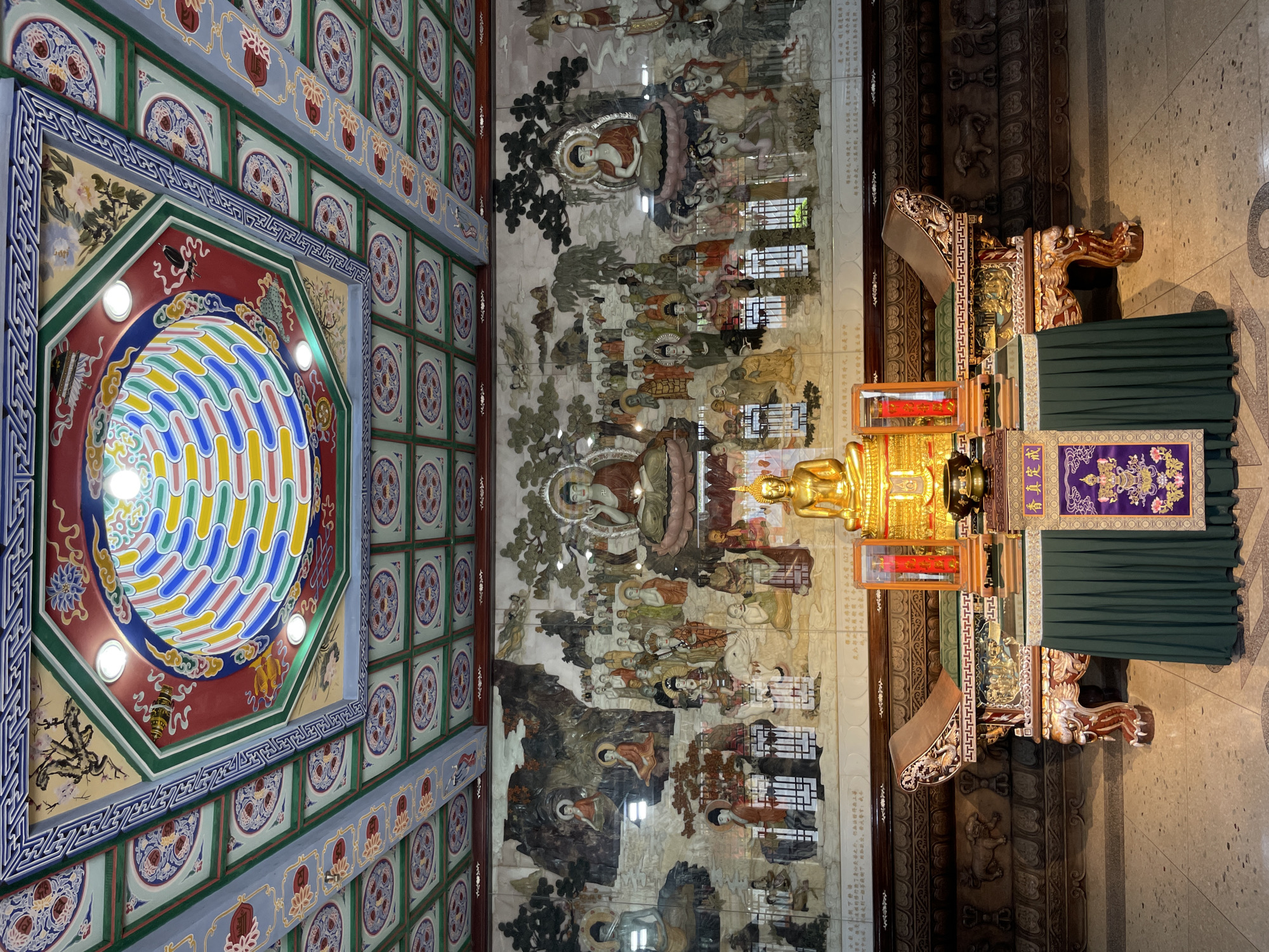 The image depicts the interior of a temple, likely located in Yanta District, China, as suggested by the architecture and religious iconography. The ceiling is elaborately decorated with an octagonal design featuring colorful geometric patterns and traditional Chinese motifs, surrounded by a grid of square panels with floral designs. A large mural adorns the wall, depicting a scene with numerous figures and Buddhas in various postures, possibly illustrating a religious narrative. In the foreground, a golden Buddha statue sits on an ornate altar, covered with a green cloth and decorated with a purple banner with Chinese characters. The altar is flanked by two intricately carved wooden chairs. The floor is made of light-colored tiles, and the lighting is bright, highlighting the intricate details of the temple's interior. The overall impression is one of ornate decoration and religious significance.