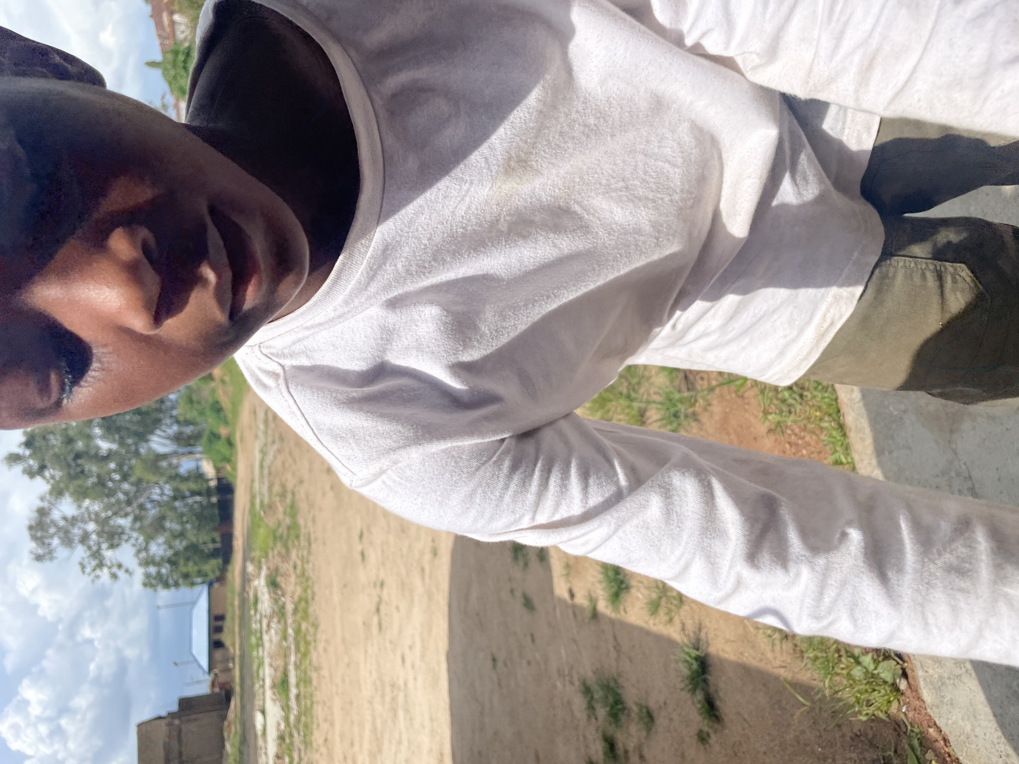 A medium close-up shot shows a person from the chest up, looking down and slightly to the right. They are wearing a white long-sleeved shirt, which has a faint yellow stain on the front. Their hands are not visible. The background is an outdoor scene in what appears to be a rural or undeveloped urban area, possibly Jos, Nigeria, with sandy ground, patchy grass, and a few buildings in the distance. One building has a blue roof, and another is a taller, unfinished structure. The sky is cloudy. The lighting suggests it is daytime.