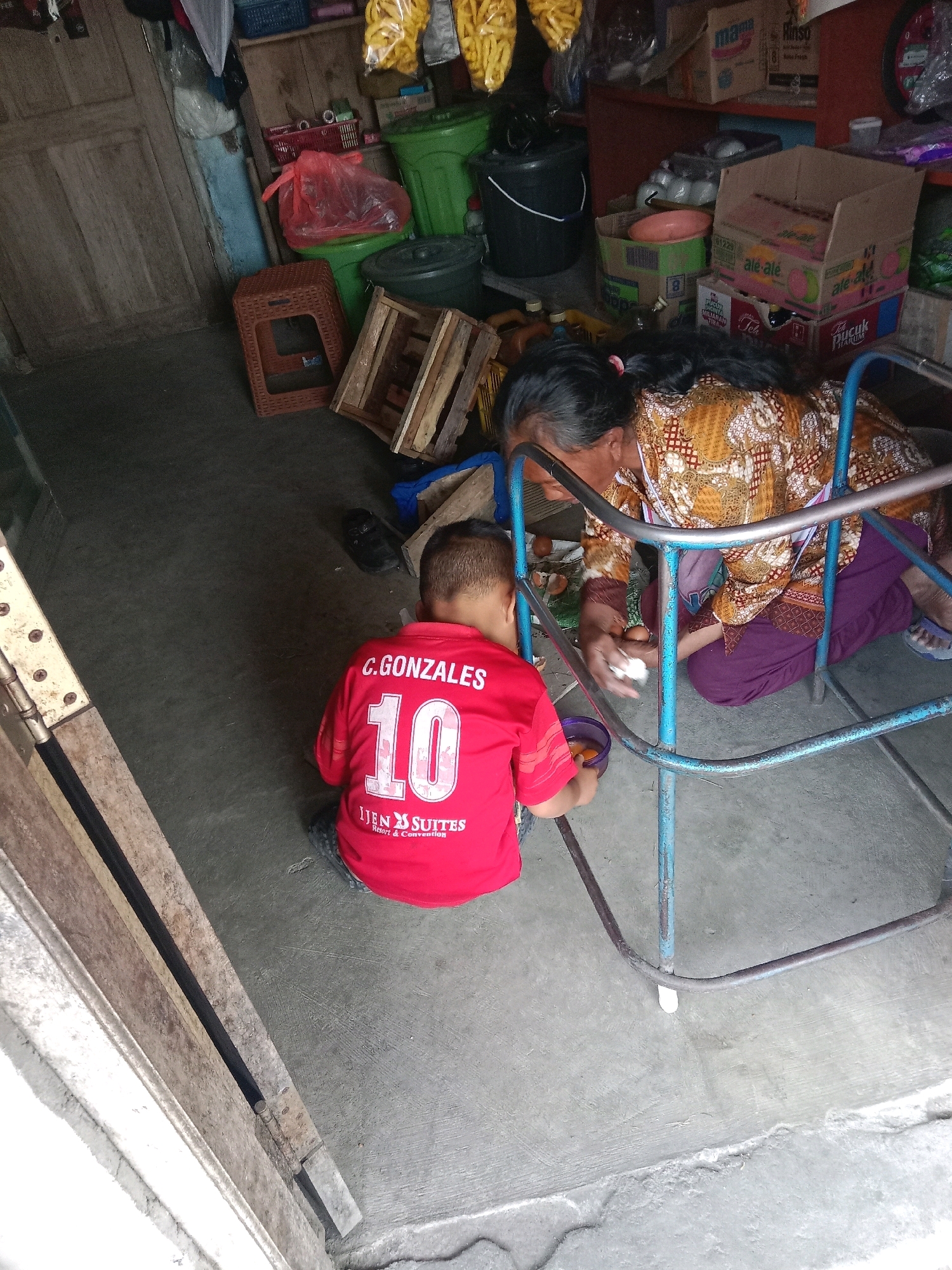 An overhead shot captures an indoor scene in Banyumudal, Indonesia. In the foreground, a young boy in a red shirt with "C. GONZALES 10" and "IJEN SUITES Resort & Convention" printed on the back is seated on the floor. He is holding a small purple bowl containing what appears to be raw egg. Facing him is an elderly woman wearing a patterned top and a purple skirt, kneeling and holding eggs in her hands. She is positioned within a blue metal walker frame. The environment is cluttered, with various objects scattered around the floor, including crates, plastic bags, and containers, suggesting a storage or workshop area. The walls and floor are made of concrete and wood.