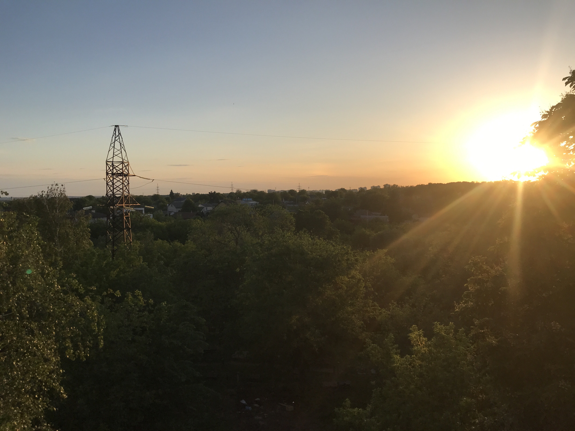 The image shows a view of Kharkiv, Ukraine at sunset. An electrical transmission tower stands to the left of the frame. A dense canopy of green trees fills the foreground and midground. Buildings and houses are visible in the background. The sun is setting on the right side of the image, partially obscured by foliage, casting bright rays across the scene. The sky is a gradient of light blue to orange.