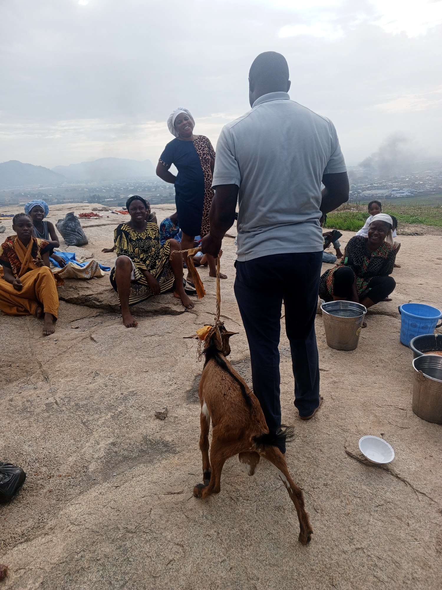 The image depicts a group of people gathered on a rocky surface, possibly a hilltop or elevated area, overlooking a city. The weather appears overcast with a cloudy sky.
In the foreground, a man stands holding a goat by a rope. Several women are seated on the ground, some smiling and observing the man and the goat. Various objects are scattered around, including buckets, a white plate, and bags. The women are wearing various dresses and headscarves. One is dressed in yellow and gold and seated next to a woman in a black and yellow dress. The other women are wearing headscarves.
In the background, there is a cityscape with buildings and what appears to be smoke rising in the distance. The location of this scene is Unknown, Unknown.