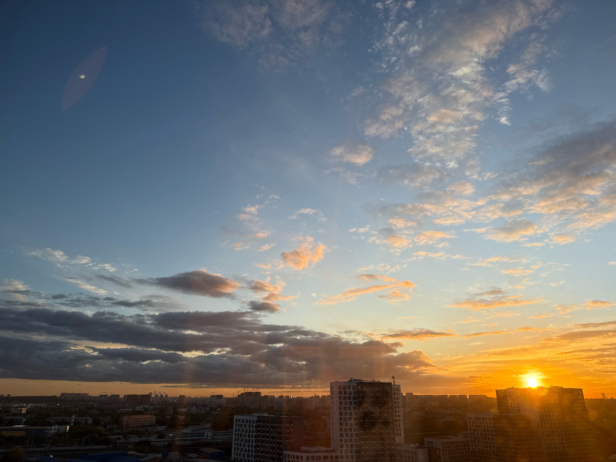 The image captures a cityscape at sunset, likely taken from an elevated perspective. A hazy, oval-shaped lens flare with a bright spot in the center is visible in the upper left portion of the sky. The sky transitions from a clear blue at the top to shades of orange and yellow near the horizon, where the sun is setting, casting warm light and long shadows. Scattered clouds, some tinged with pink and orange, dot the sky. In the foreground, a dense urban environment with numerous buildings, including high-rises and lower structures, stretches across the landscape. Some buildings appear to be under construction, indicated by visible cranes. The city appears to be located in Lyubertsy, Russia, as suggested by the architectural style and urban layout. No people are discernibly present in the image. The overall impression is one of an urban sunset, with the city bathed in the warm glow of the setting sun.