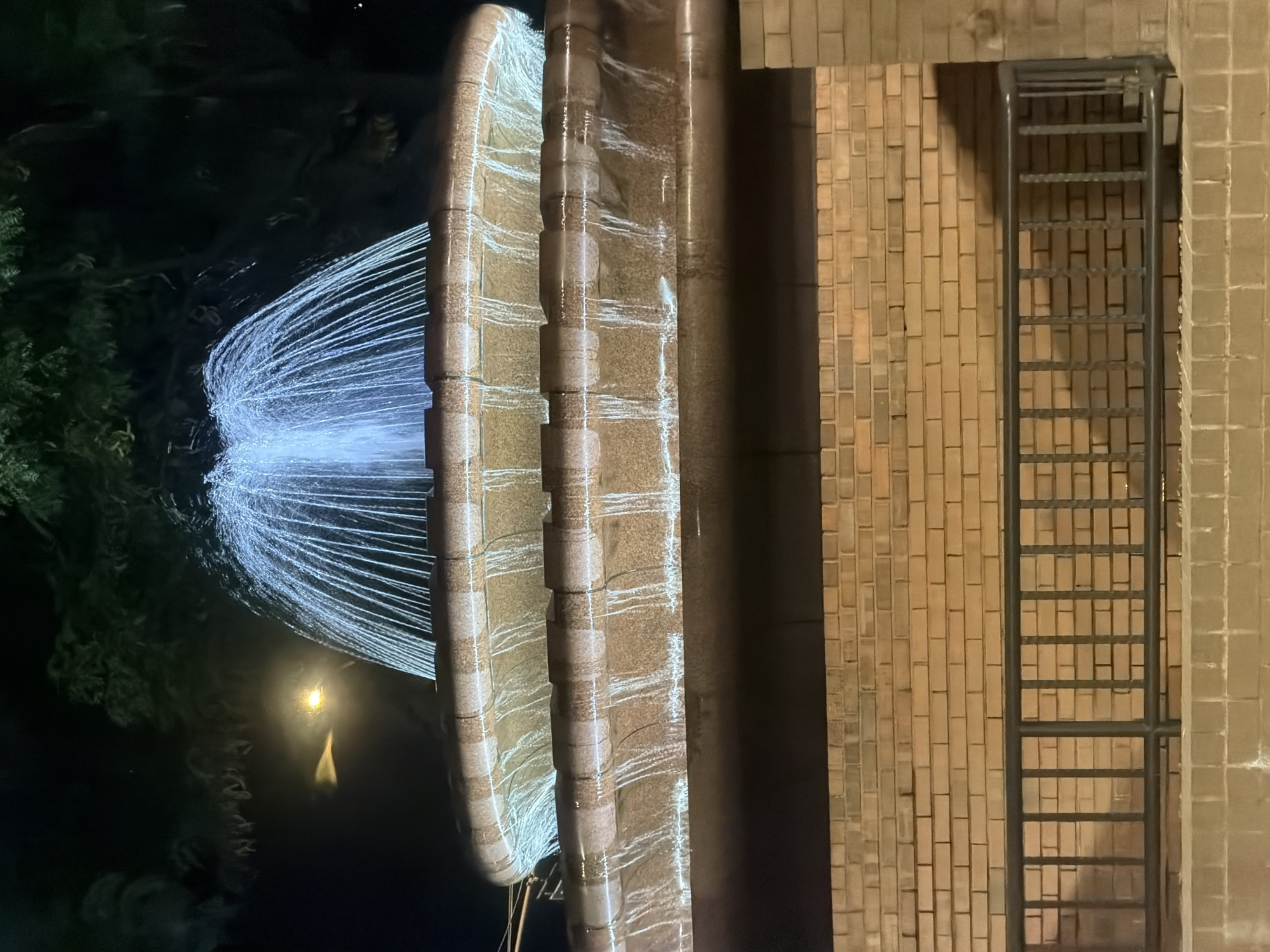 A multi-tiered fountain is illuminated by artificial lights at night. The fountain has water spraying upwards in a fan shape from the top tier, and cascading down the sides of the tiers. The fountain structure is made of textured stone with darker, speckled accents. Below the fountain, a brick wall serves as a backdrop. In front of the wall, a dark metal railing with vertical bars runs horizontally, separating the viewer from the fountain area. The scene is set in a dark outdoor environment, likely a park or public square, with hints of trees in the background. No people are visible in the image. The lighting suggests it is nighttime. The location is not explicitly identifiable as New York City.