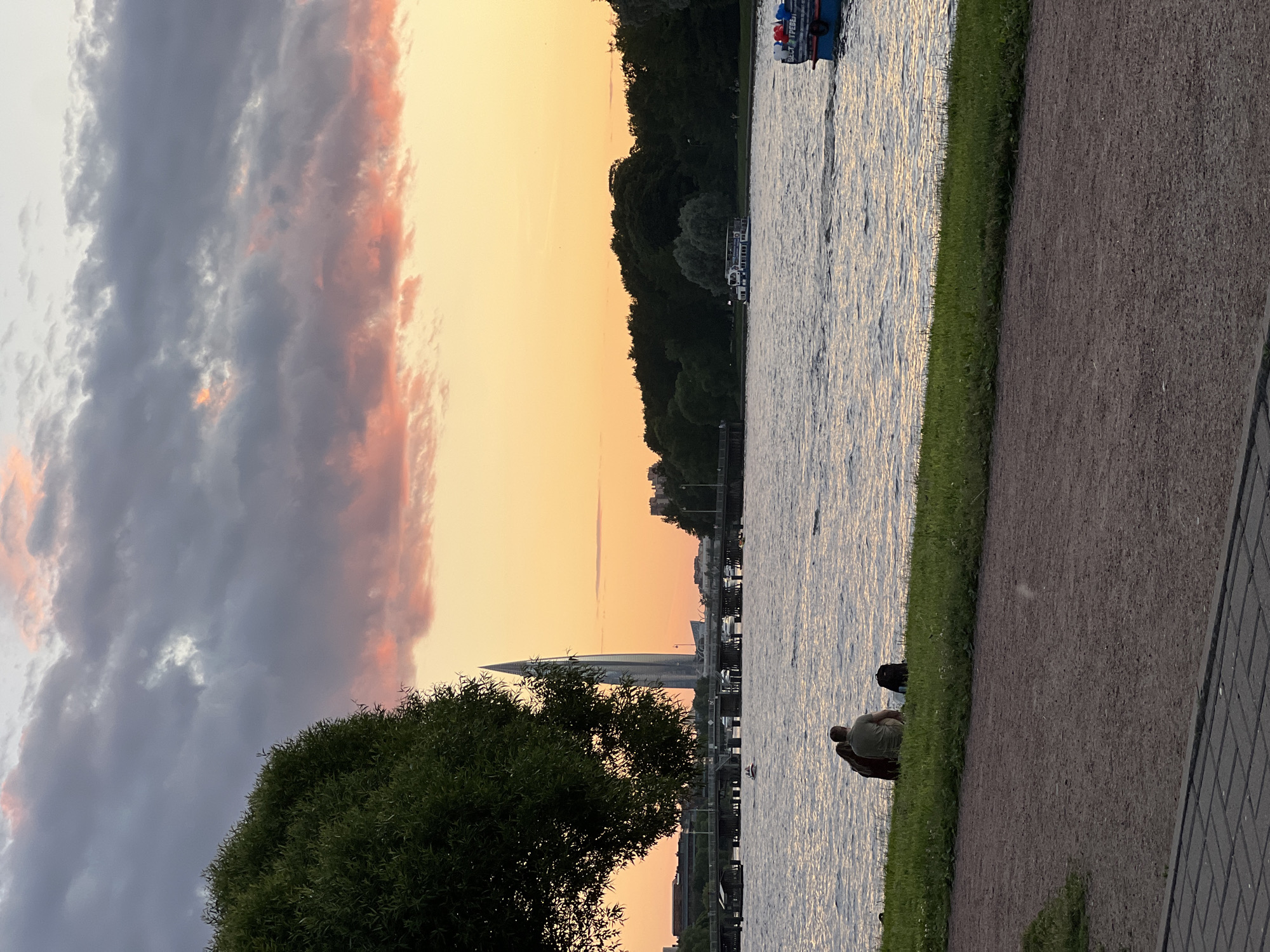 The image depicts a sunset scene over a body of water, likely a river or canal, in Saint Petersburg, Russia. In the foreground, a pathway made of gravel and paving stones leads to a grassy embankment where two individuals are seated facing the water, with a backpack beside them. A large tree with dense foliage partially obscures the left side of the frame.
In the mid-ground, the water is rippled, reflecting the soft light of the sky. A bridge crosses the water in the distance, supported by numerous wooden pilings. Several boats are visible on the water, including a larger passenger boat moving from left to right.
The background features a skyline dominated by a distinctive, tall, slender skyscraper with a pointed top, likely the Lakhta Center, a prominent landmark in Saint Petersburg. The sky is filled with a gradient of colors, transitioning from a pale yellow near the horizon to pink and gray clouds higher up. The overall atmosphere is serene, with the setting sun casting a warm glow over the urban landscape.