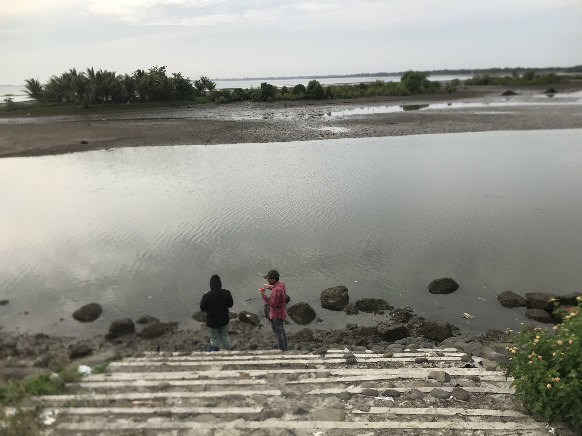 Two individuals are standing on the muddy bank of a body of water. The person on the left is wearing a black hooded sweatshirt and light-colored pants, facing away from the camera. The person on the right is wearing a red plaid shirt, dark pants, and a baseball cap, facing left and appearing to hold a small object. They are positioned near a cluster of rocks. In the foreground, a set of concrete stairs with stones embedded in them leads down towards the water. The background shows a stretch of muddy land with scattered vegetation, including palm trees, and a distant shoreline with more trees under an overcast sky. This scene is likely located in the Lhokseumawe, Indonesia area, potentially near a coastal or estuarine environment.