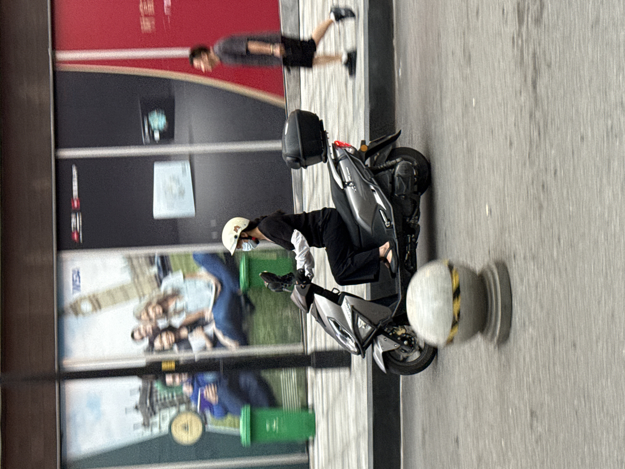In Yuzhong District, China, a person wearing a white helmet and a mask rides a silver scooter. The scooter has a black storage box on the back and is in motion on a street. In the background, there are large advertisements displayed on buildings. One advertisement features images of people and a clock tower. Another advertisement is a solid red panel. To the right of the scooter, a person in a grey t-shirt and black shorts is walking away from the camera. Two green trash cans are visible in the mid-ground behind the scooter. A blurry grey spherical object with yellow and black stripes is in the foreground. The street surface is grey and appears wet in some areas.