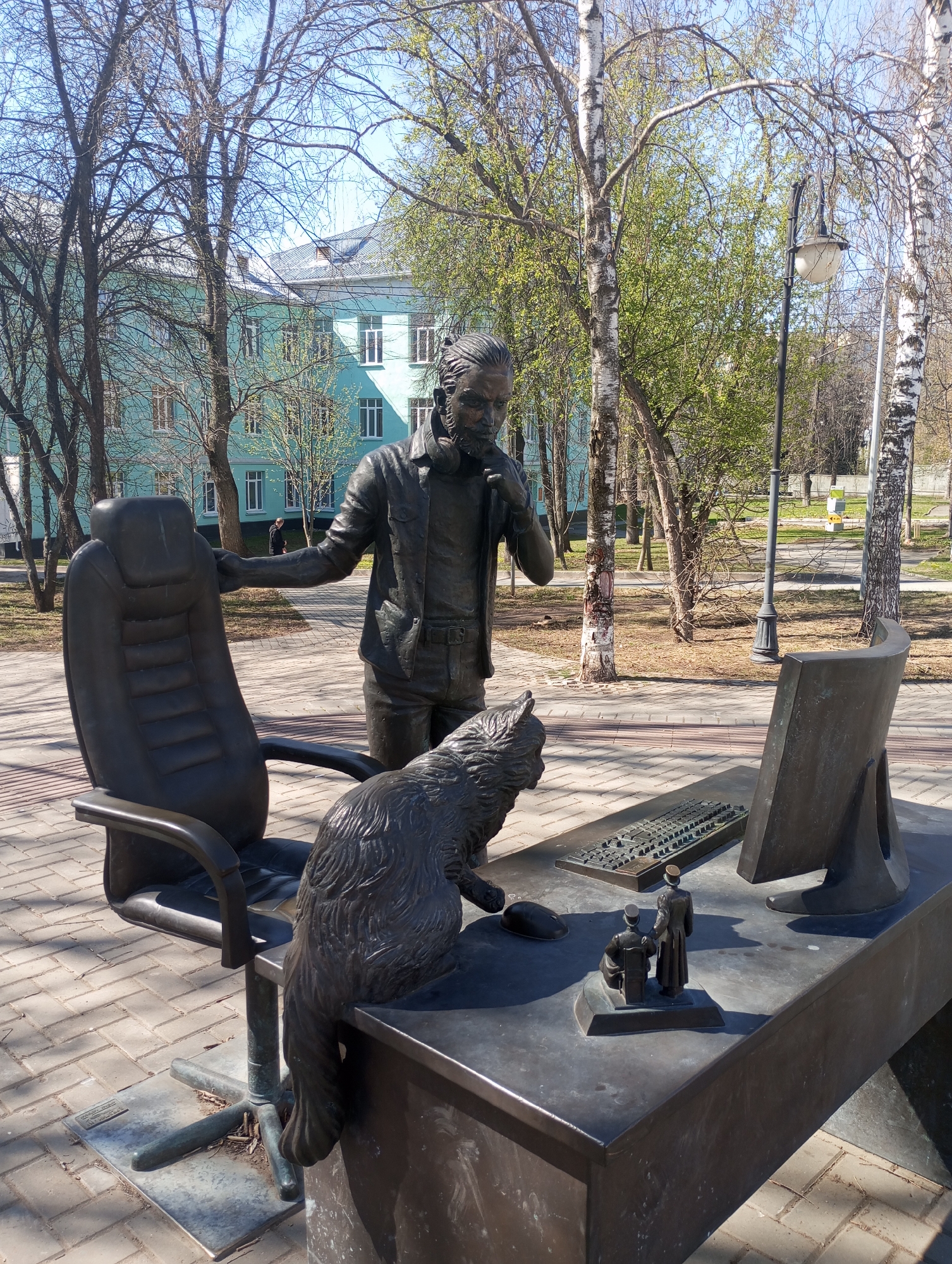 The image captures a bronze statue in Pervomaysky, Russia. The statue depicts a man standing beside an office desk and chair, with a cat sitting on the desk. The man is dressed in a casual outfit, and has his hand near his face, as if in thought. The cat is positioned close to a miniature computer mouse. The desk also features a keyboard and a computer screen, and two small human figurines. In the background, there's a light-blue building and several trees. The scene is set on a paved area, suggesting a park or public space.
