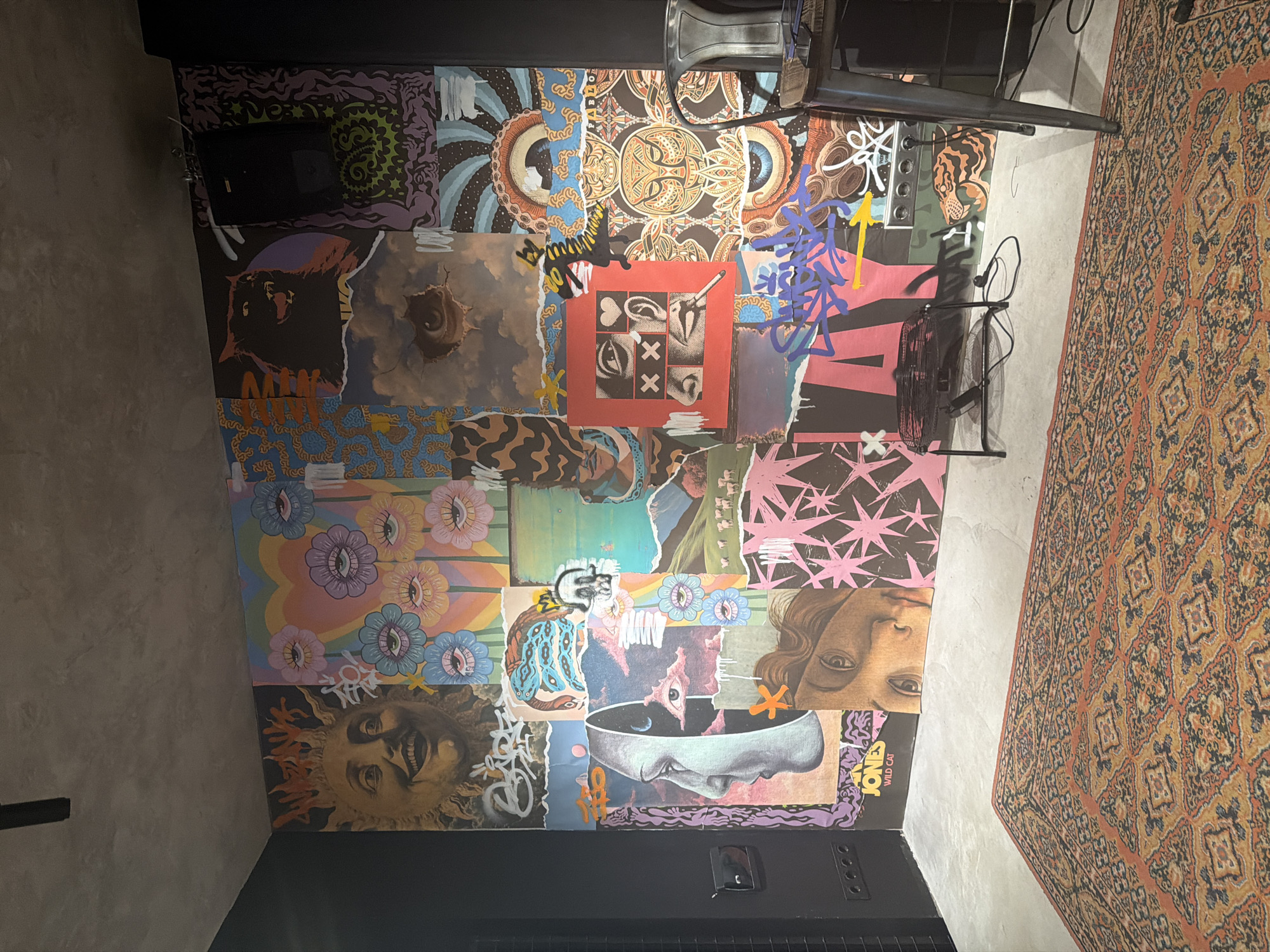 The image is a high-angle shot of a room featuring collage artwork on a wall. The collage is composed of numerous smaller images pieced together, displaying various motifs including faces, eyes, flowers, and abstract patterns. The color palette is diverse, ranging from muted earth tones to vibrant blues, pinks, and purples. The bottom edge of the wall where the collage ends is a dark, possibly black, painted section. In the upper right corner, there is an area of decorative orange, brown, and black patterned rug. A metal folding chair is in the upper part of the frame, and a black electrical fan is next to the folding chair. The left side of the image shows a gray textured wall that extends into the background. The scene is indoors with no people visible. The image does not contain any visual information that confirms its location as Naberezhnye Chelny, Russia.