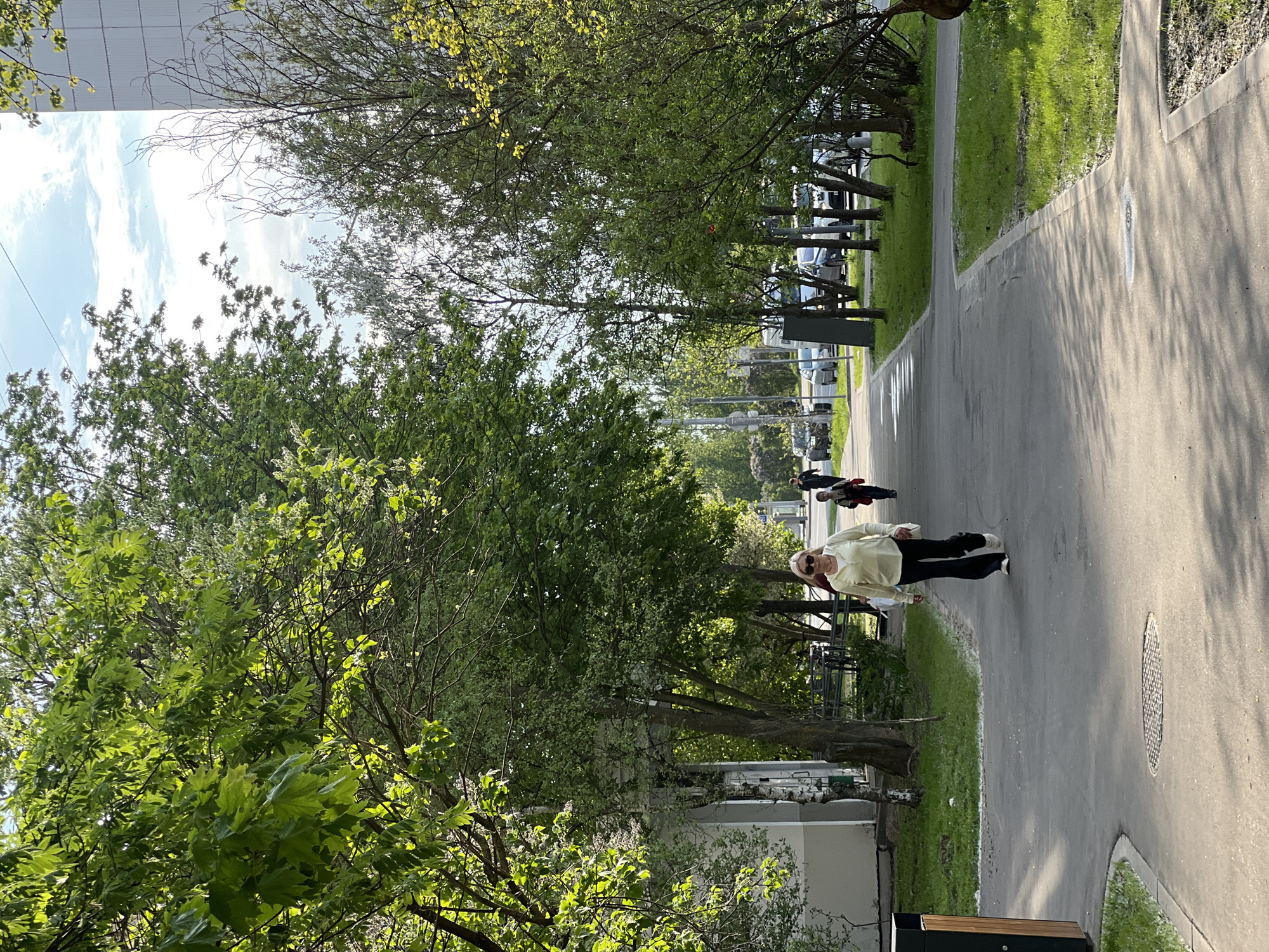 Here is a detailed description of the image:
**Scene Overview:**
The image presents an outdoor scene along a paved walkway adjacent to a green space filled with trees. Sunlight casts shadows across the pavement. The photo seems to be taken in a city environment, lined with apartment buildings and vehicular traffic in the background.
**Subjects and People:**
There is a woman wearing a light-colored top, dark pants, sunglasses, and white sneakers walking along the walkway. Behind her, there are two additional people. One is wearing red top, and the other is wearing black. They appear to be traveling in the same direction.
**Objects:**
* A grey pavement/sidewalk.
* Trees line the side of the pathway.
* Apartment buildings.
* Vehicles are parked along the side of the road in the distance.
* A black trash can is present in the lower part of the image.
**Actions and Interactions:**
The primary action is people walking along the path. The woman in the foreground walks at a pace that suggests exercise.
**Environment:**
* The scene is outdoors during the day, likely in the late morning or early afternoon based on the shadows.
* The trees indicate an urban park or green space is present alongside the walkway.
**Location Estimate:**
Based on the architectural style of the apartment buildings and the overall urban layout, the image shows the city of Moscow, Russia.