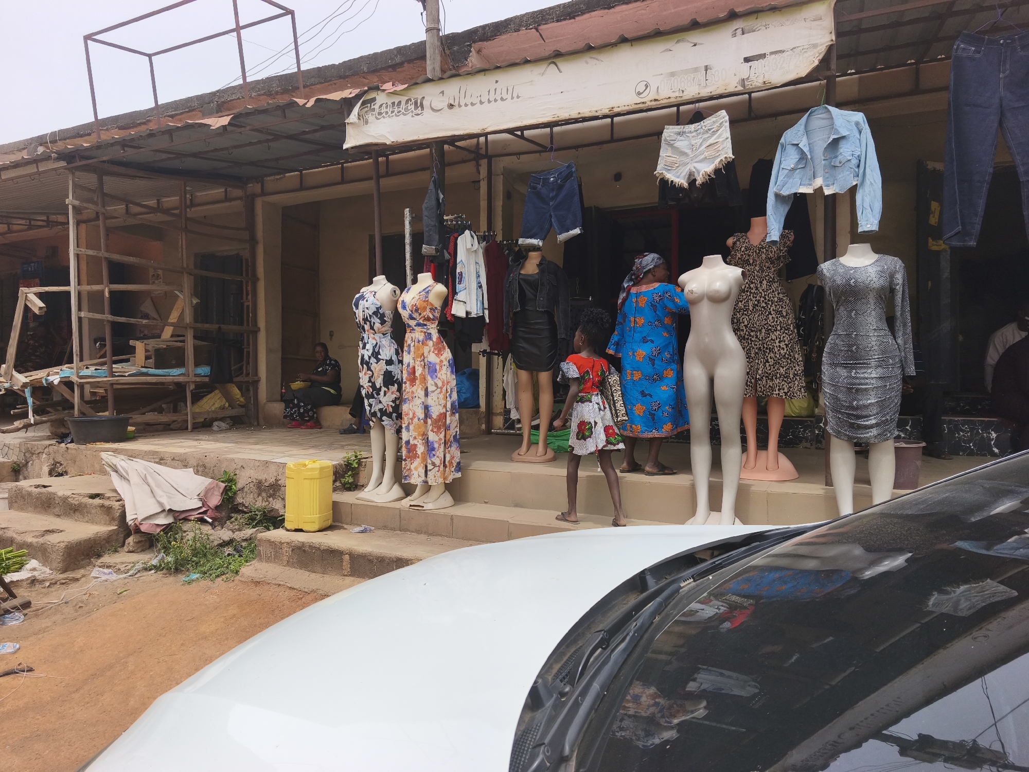 The image captures an outdoor clothing shop in Jos, Nigeria, named "Fancy Collection." The shop has a corrugated metal awning extending over its entrance, beneath which a white sign displays the shop's name in a decorative font, with a partially visible phone number to the right.
In front of the shop, several mannequins display a variety of women's clothing, including floral patterned dresses, a leopard print dress, a snake print dress, and a blue patterned dress. Above the mannequins, denim shorts, jeans, a light blue denim jacket, and a black jacket are hung for display.
A set of worn concrete steps leads up to the shop's entrance from a reddish, unpaved dirt area. A yellow plastic jerrycan sits on one of the steps, and a folded umbrella lies near the bottom amidst some weeds and litter. To the left of the main shop, a rudimentary wooden scaffolding structure is visible, with a black bucket on the ground.
Several people are present: a small child, likely a girl, wearing a colorful patterned dress, stands near the mannequins. An adult woman sits on the steps to the far left, while another woman, wearing a blue patterned dress similar to one on a mannequin, is partially visible inside the shop. Other indistinct figures can be seen in the background within the shop.
The scene appears to be taking place during the daytime under an overcast or hazy sky, as suggested by the diffused light and lack of strong shadows. The foreground is partially obscured by the hood and windshield of a white car, reflecting parts of the street scene. The overall setting reflects a common commercial environment in an urban or semi-urban area.