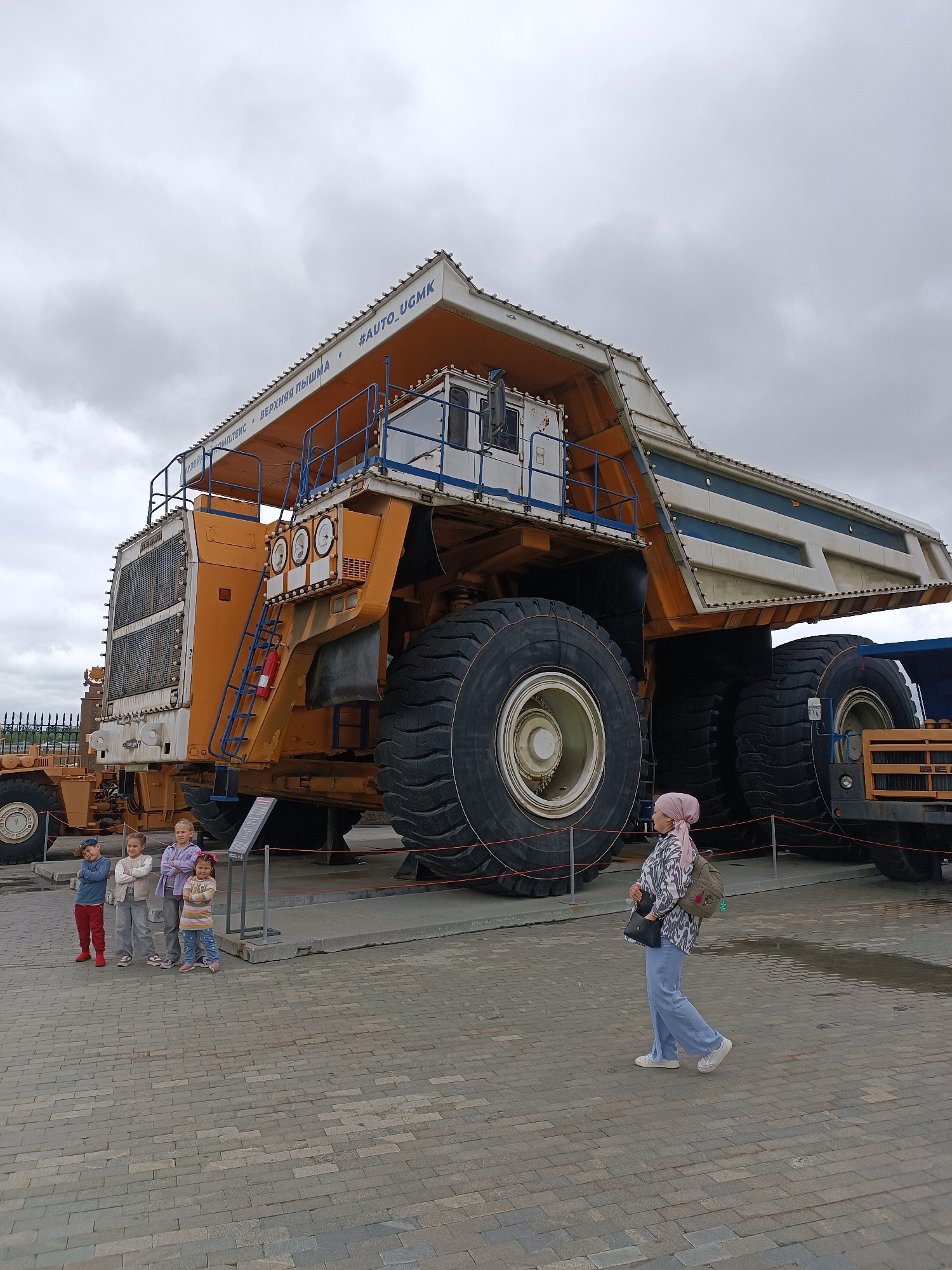 In Verkhnyaya Pyshma, Russia, a colossal yellow mining truck, identified by the text "#AUTO_UGMK" and "МУЗЕЙНЫЙ КОМПЛЕКС • ВЕРХНЯЯ ПЫШМА" on its upper structure, stands as a prominent exhibit. Four children, two boys and two girls, are positioned in the foreground, facing the truck with their arms crossed. To the right of the children, a woman wearing a pink hijab, a patterned shirt, and light blue pants walks away from the viewer, carrying a backpack and a purse. The ground is paved with gray brickwork, and a low barrier line separates the exhibit from the viewing area. The sky is overcast with gray clouds.