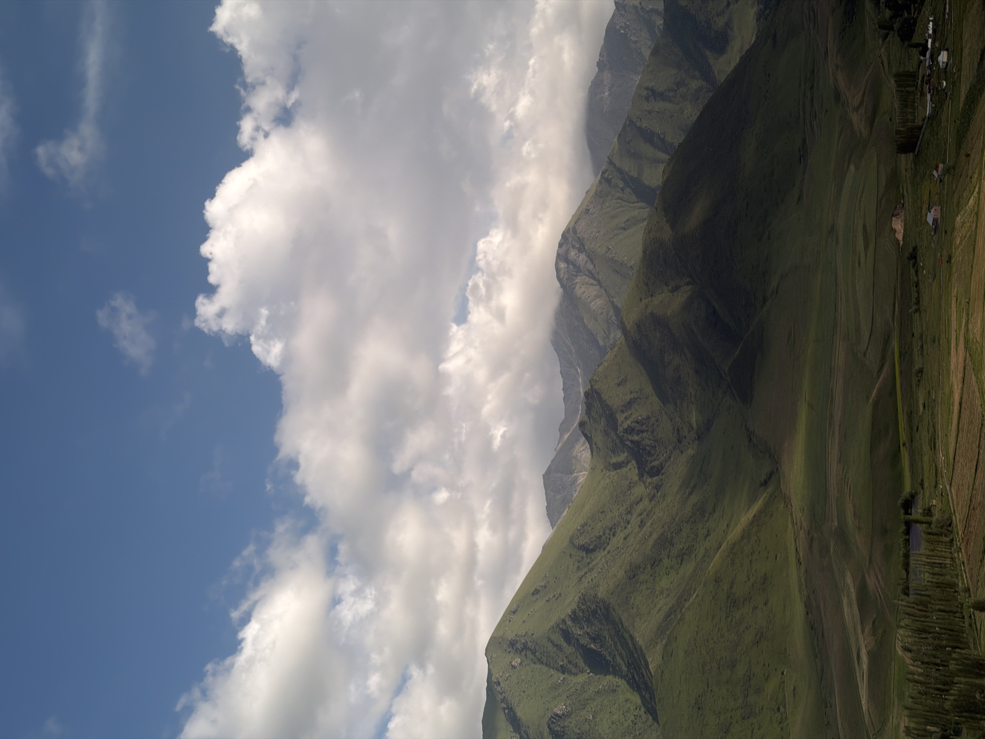 This media file captures a dramatic mountain landscape, likely in Karagai Bulak, Kyrgyzstan. The scene is divided by a clear diagonal line, with the left side dominated by a vibrant blue sky filled with fluffy white clouds. The right side features imposing, verdant mountains that rise steeply. These mountains are covered in lush green vegetation, with visible terraced fields and a small cluster of houses suggesting human habitation and agricultural activity. The terrain appears rugged and uneven, with deep ravines and sharp ridges.
The sunlight suggests it is daytime, likely late morning or early afternoon, given the brightness of the sky and the shadows cast on the mountains. The weather appears fair, with a mix of sun and clouds. The overall mood is one of natural grandeur and perhaps a sense of isolation due to the remote mountainous setting. No people are visible, nor are there any distinct objects that draw attention apart from the natural elements and the distant dwellings. There is no visible text in the image.
