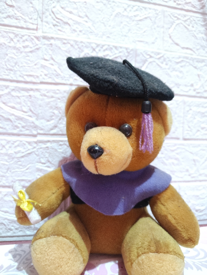 The scene, located in Astanajapura, Indonesia, presents a close-up, eye-level view of a brown plush teddy bear. The teddy bear is seated and adorned in miniature graduation attire, consisting of a black felt mortarboard with a black cord and a purple tassel positioned atop its head, and a purple felt stole draped over its shoulders, covering a small visible section of a black garment. The bear has two small, round, black plastic eyes and a black plastic nose. In its left arm, it holds a small, white scroll, resembling a diploma, secured with a yellow ribbon. The background features a textured white wall with a distinct, raised, brick-like pattern. The bear rests on a light-colored surface with a faint, repeating pattern, which is partially visible at the bottom of the frame. No human subjects are present.