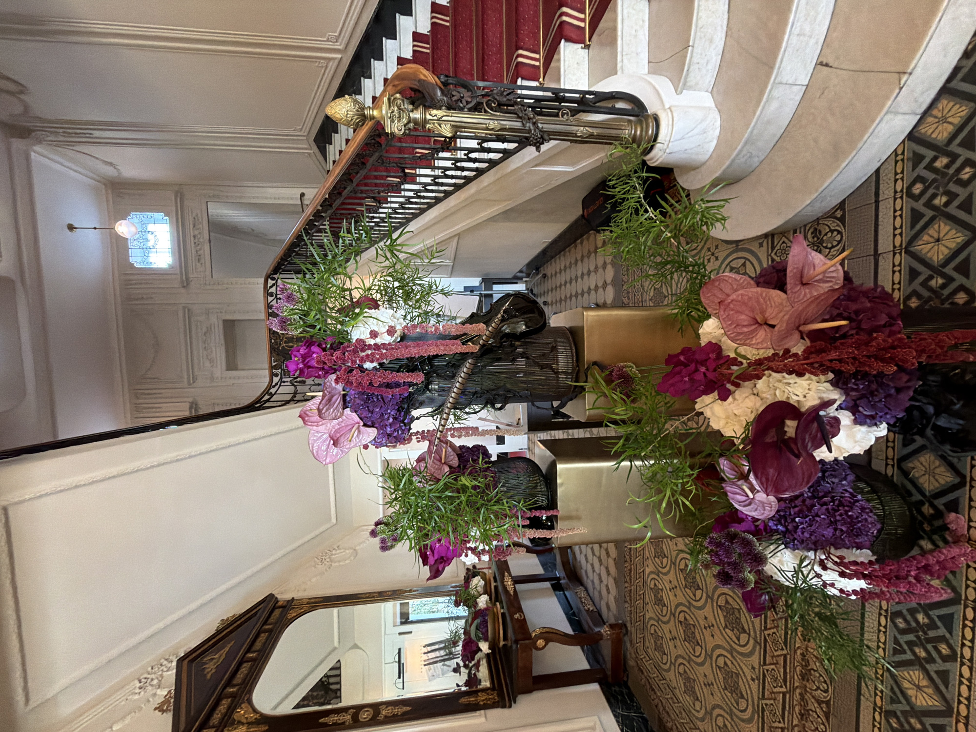 The image depicts an ornate interior hallway with a grand staircase. The staircase has a red carpet runner with two gold stripes, and is flanked by a decorative black metal railing with gold accents. The walls are cream-colored and feature elaborate molding. A large ornate mirror with a gold frame hangs on the left wall above a dark wood console table. In the foreground, several large floral arrangements are displayed. These arrangements feature a mix of purple, pink, and white flowers, including anthuriums and hydrangeas, with cascading red amaranth and green foliage. The floor is covered in a patterned tile with intricate geometric designs. A hanging light fixture with a frosted glass shade is visible on the ceiling. The scene suggests a luxurious and well-decorated interior space, possibly a hotel lobby or a grand residence.