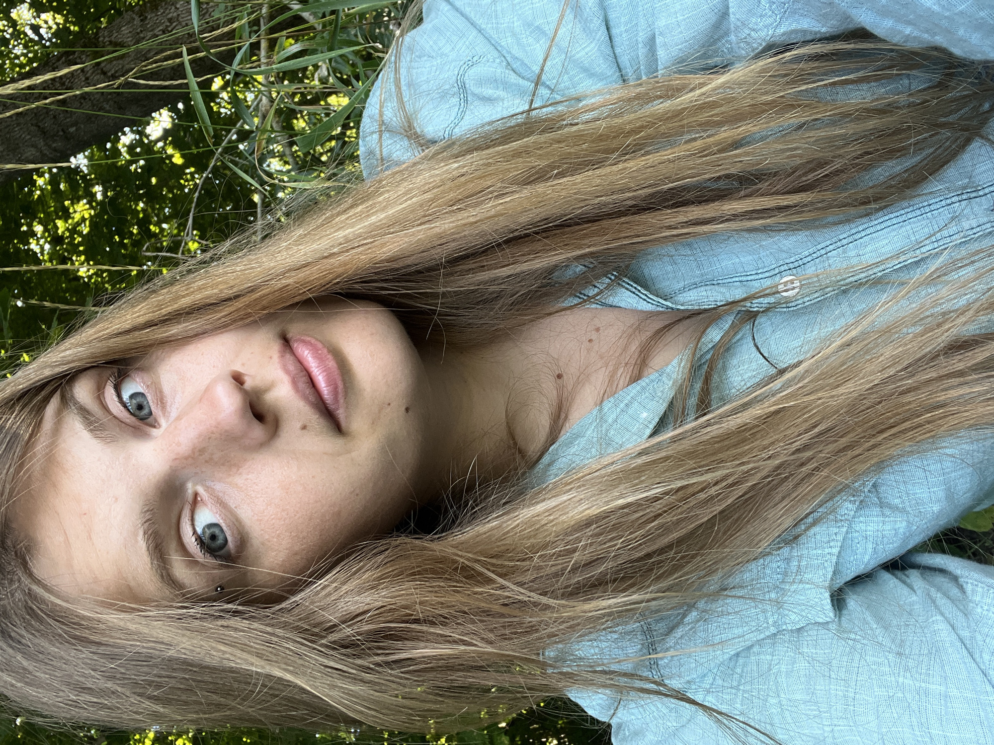 A young woman with long, blonde hair and blue eyes is pictured in an outdoor setting. She is wearing a light blue collared shirt with buttons. Her hair is long and falls over her shoulders, partially obscuring her face and the shirt. She has a piercing above her left eyebrow. The background consists of lush green foliage and trees, suggesting a natural environment. The lighting indicates it is daytime, with sunlight filtering through the leaves. The woman is looking directly at the camera with a neutral expression. The image is a close-up shot, focusing primarily on the woman's face and upper body. The location is identified as Германавіцкі сельскі Савет, Belarus.