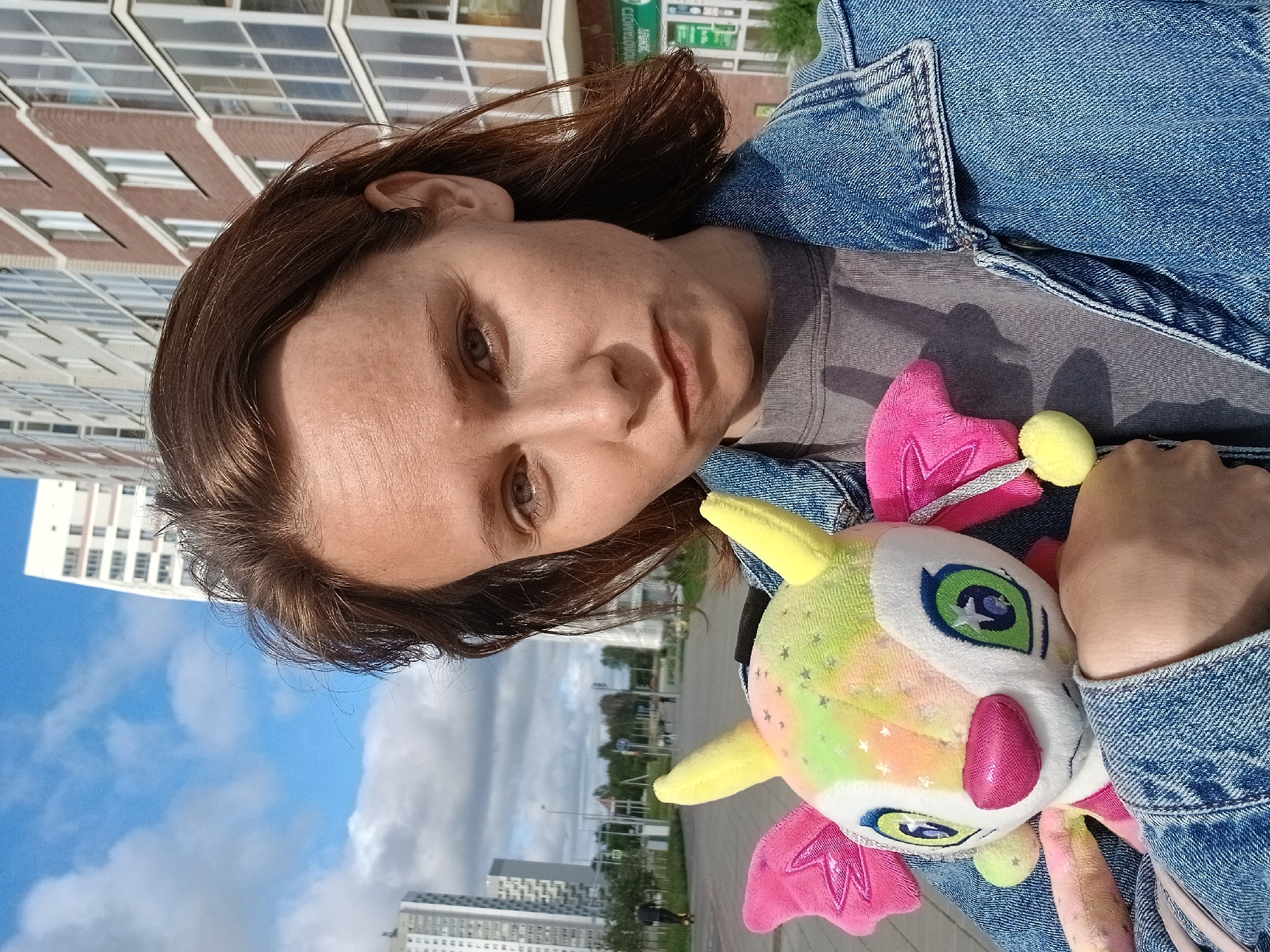 In Yekaterinburg, Russia, a young woman with brown hair and light skin, wearing a denim jacket and a grey t-shirt, holds a colorful plush toy with yellow ears, pink wings, and green eyes. She is positioned outdoors with a street and modern buildings in the background. The sky is partly cloudy, and the lighting suggests it is daytime.