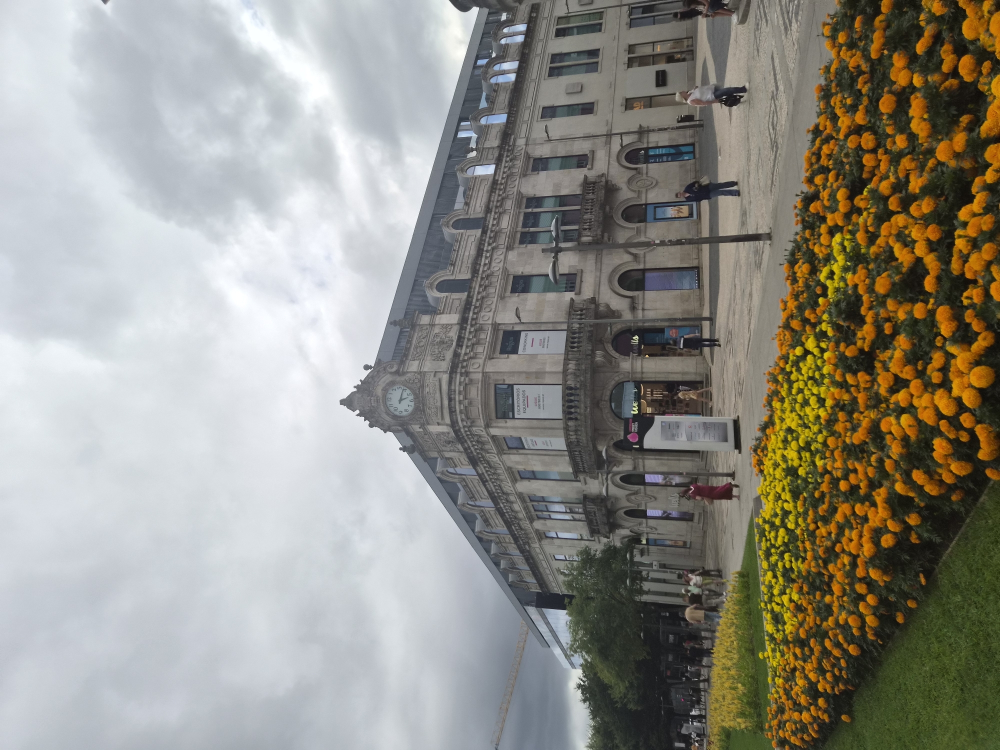 The image captures a view of a grand, ornate building with a clock tower, situated in Braga, Portugal. The architecture of the building is classical, featuring stone facades, arched windows, and decorative elements. A modern glass-paneled extension is visible to the left, contrasting with the older structure. In the foreground, a vibrant flower bed with numerous orange and yellow marigolds creates a striking visual. Several people are present on the paved area in front of the building, walking and interacting, though none are in the immediate foreground. The sky is overcast with dense clouds. A construction crane is visible in the distance, behind the trees on the left side of the image. The overall scene depicts a bustling urban environment with a blend of historical and contemporary architecture.