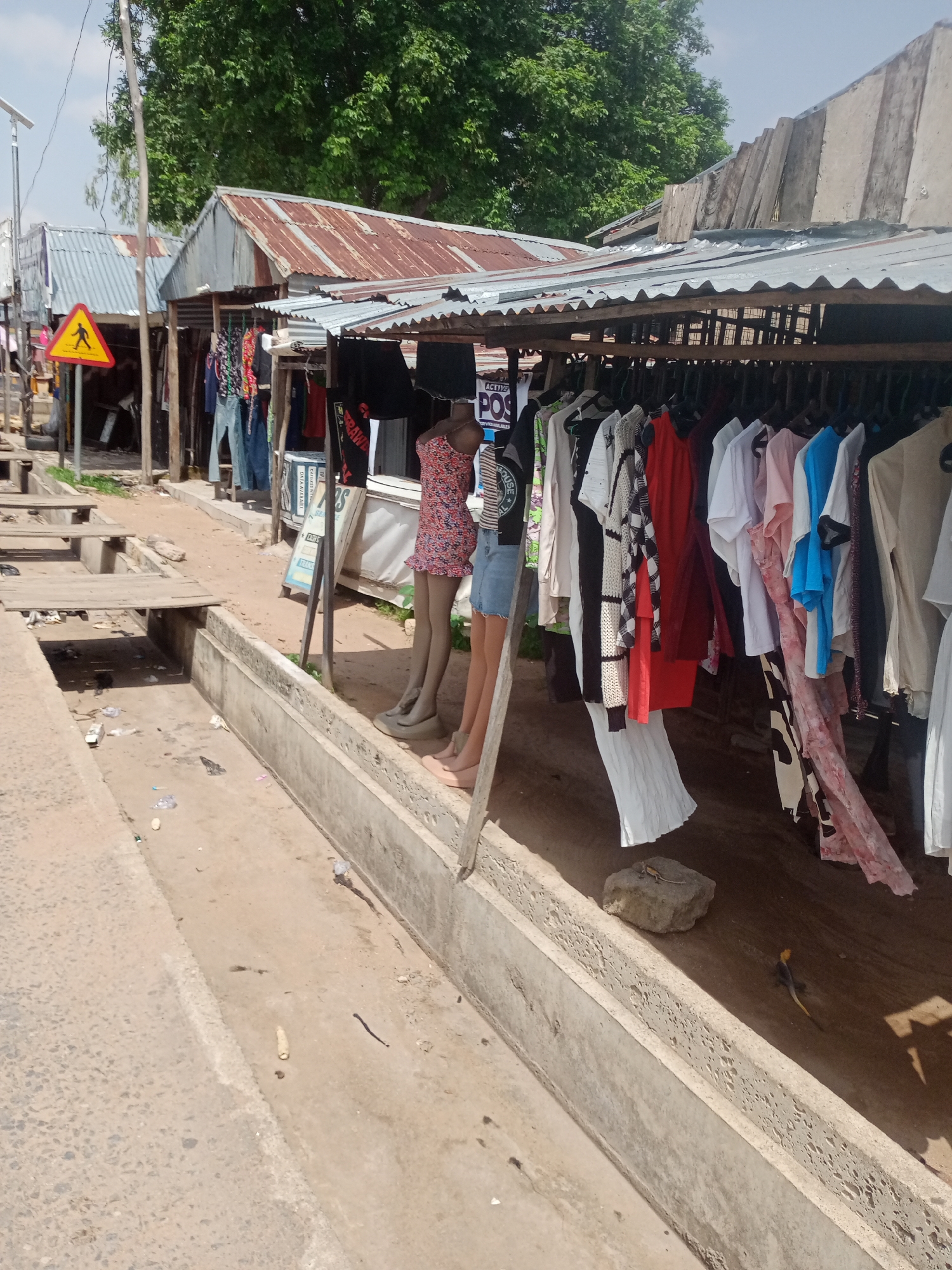 The image depicts an outdoor market scene in Jalingo, Nigeria. In the foreground, a concrete drainage channel runs alongside a dirt path. Several pieces of trash are scattered on the ground.
The main subjects are a row of market stalls with corrugated metal roofs, some of which appear weathered. Clothes are displayed for sale on hangers hanging from wooden poles. Among the clothing items are dresses, shirts, and skirts in various colors and patterns. Two mannequins are partially visible, one wearing a floral print dress and the other a denim skirt, indicating that the stalls are selling apparel.
In the background, more market stalls and structures are visible, along with dense green trees. A yellow triangular warning sign with a black silhouette of a person walking is mounted on a pole to the left of the stalls. The sky is bright and appears to be a clear day, suggesting it is daytime. The overall scene conveys a sense of everyday commerce in a bustling open-air market.
There is some visible text on a sign attached to the stall, partially obscured, reading "POS". Another sign with text is also visible behind the mannequins. Two small lizards are present on the ground near the drainage channel, one a rock lizard and the other a agama lizard, adding a touch of local wildlife to the scene.