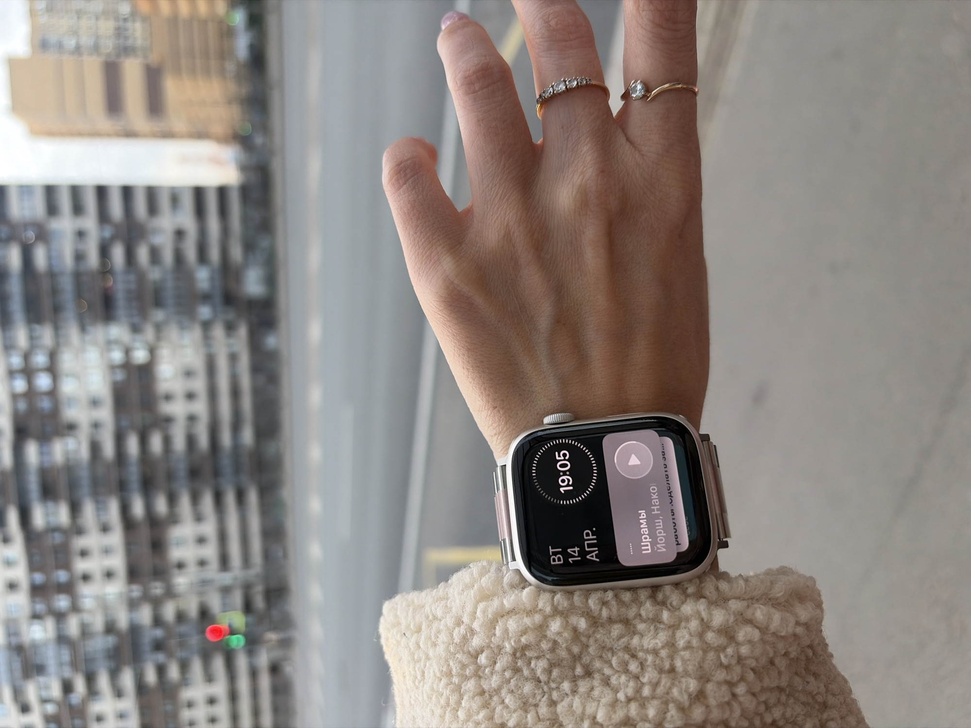 A person's hand and wrist wearing an Apple Watch is positioned in front of a window overlooking a city. The Apple Watch displays a black screen with white text and graphics; the time is 19:05. The hand wears two gold rings, one with multiple small diamonds and another with a single diamond. The wrist rests on a cream-colored, textured garment, likely a sweater or jacket. Outside the window, tall buildings with gray facades and numerous windows are visible, suggesting an urban setting. Based on the architecture and urban landscape, the location is identified as Saint Petersburg, Russia.
