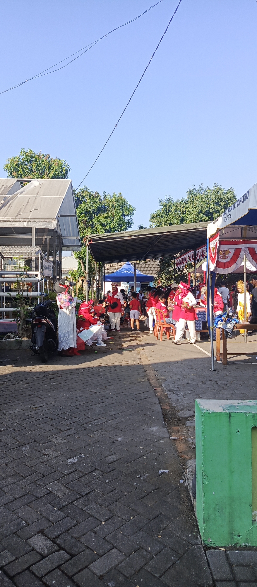 The image depicts an outdoor scene in Malang, Indonesia, likely during an event or festival. In the foreground, a paved walkway made of interlocking bricks leads into the scene. To the left, a motorcycle is parked near a building with a translucent, sloped roof. A woman in a long white dress and a wide-brimmed hat stands beside the motorcycle, facing away from the camera.
The midground is populated by a gathering of people, many wearing red and white attire, suggesting participation in a cultural event or parade. Some individuals are seated on chairs or on the ground, while others are standing. Tents and temporary structures are set up, providing shade. One tent on the right has a banner with Indonesian flag colors and text.
In the background, lush green trees are visible against a clear blue sky. The overall atmosphere appears to be one of community gathering and celebration under bright sunlight. The presence of multiple tents and decorated stalls suggests a marketplace or performance area.