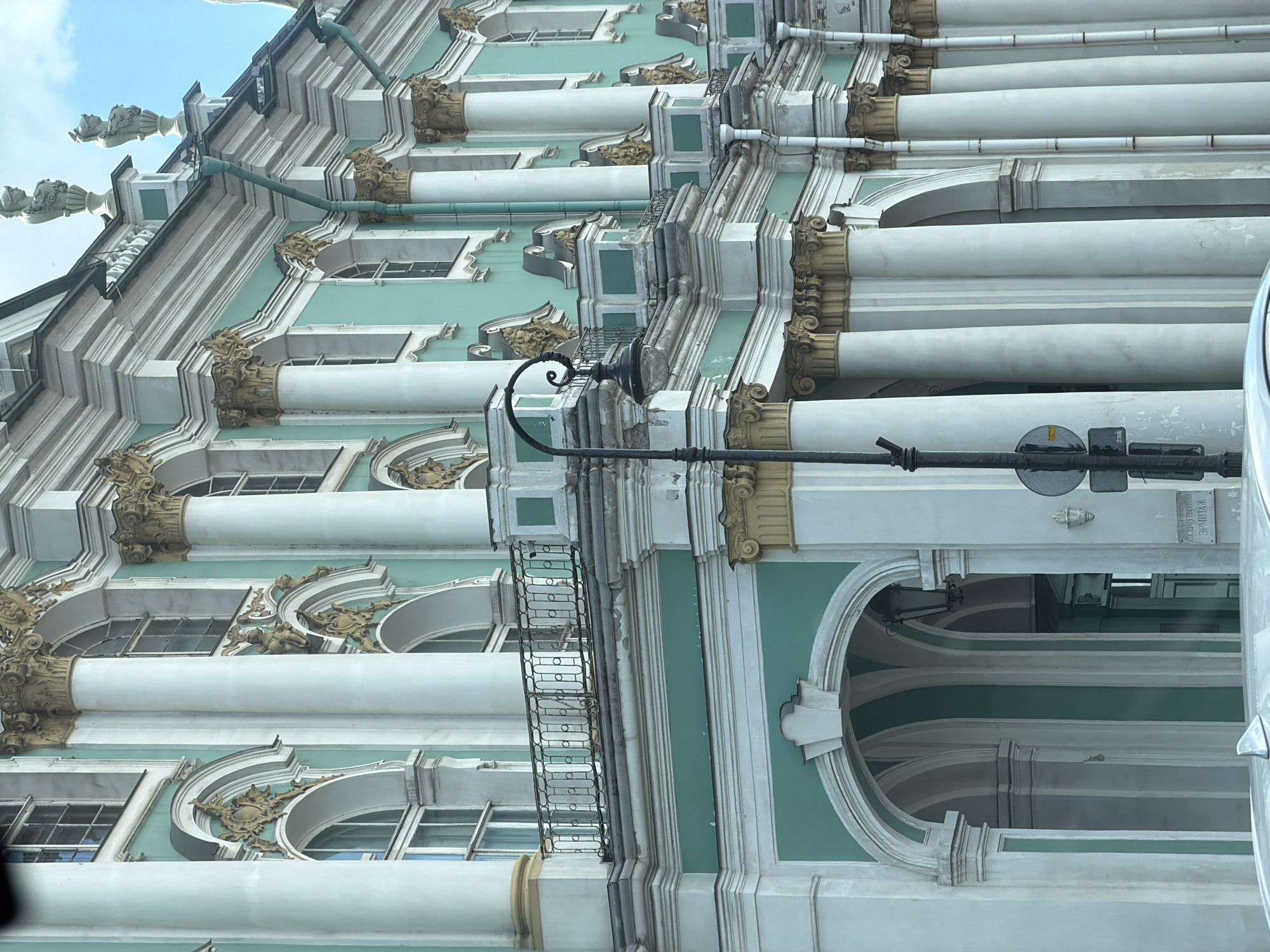 The image shows a section of the Hermitage Museum in Saint Petersburg, Russia. The building features a neoclassical architectural style with prominent white columns, ornate gold detailing, and a light teal facade. Several arched windows and a balcony with a black metal railing are visible. A streetlamp stands in the foreground, and part of a white car is in the lower left corner of the frame. The sky is visible in the upper right, with some clouds present. The scene is captured from a low angle, looking upwards at the building's facade.
