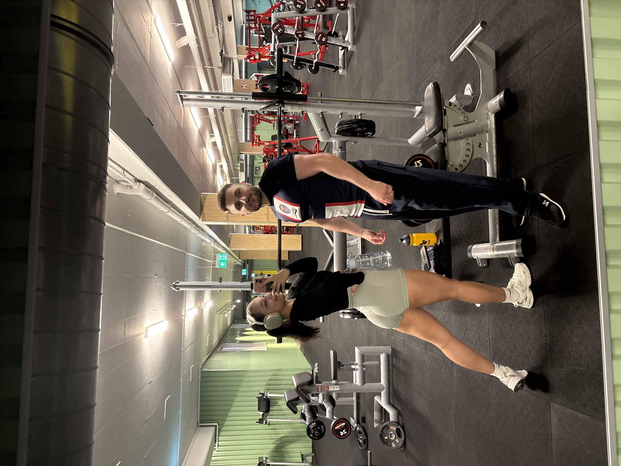 Two individuals, a male and a female, are posing for a mirror selfie in a gym. The female, on the left, is wearing a black long-sleeved shirt, light green shorts, white socks, and white athletic shoes. She has headphones on and is holding a pink smartphone, taking the picture. The male, on the right, is wearing a navy blue t-shirt with white and red accents, navy blue athletic pants, and black athletic shoes. He is standing next to a weight rack. The gym environment features various weightlifting equipment, including weight racks, benches, and free weights. The walls are painted green and white, and fluorescent lights are visible on the ceiling. The overall setting suggests a fitness facility. The location is identified as a gym in Mönchengladbach, Germany.