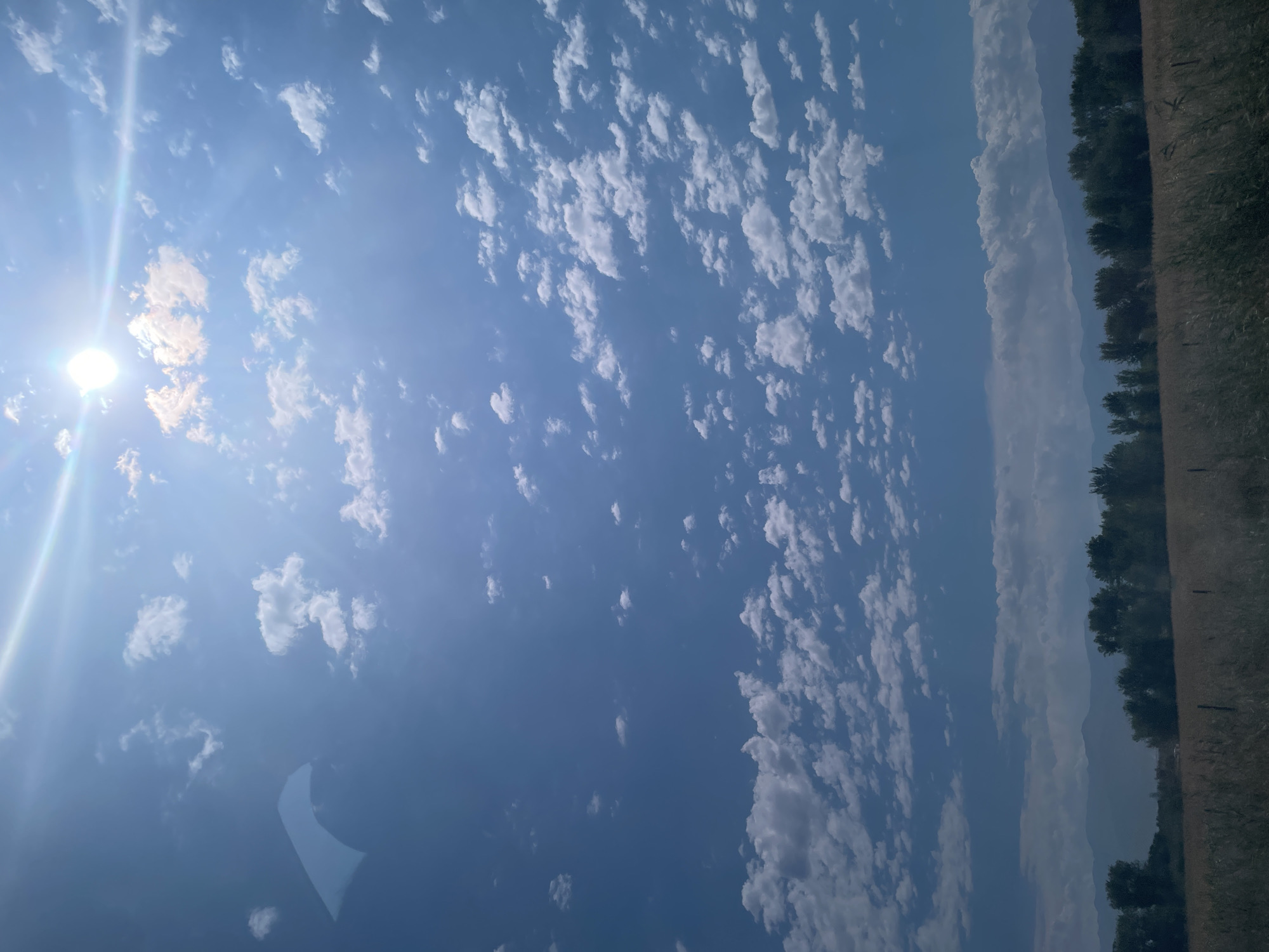This is a wide-angle, full-frame, eye-level shot of a blue sky with scattered white clouds on a sunny day. The sun is visible in the upper right quadrant, casting a bright glare and lens flare across the sky. There is a light blue, somewhat distorted, kite-like object in the upper left quadrant of the image. In the lower third of the image, a distant mountain range is visible, silhouetted against the sky. In front of the mountains, there is a line of dark green trees, and in the foreground, there is a field of dry, light brown grass with some out-of-focus fencing visible. The image appears to be taken outdoors in a rural or semi-rural area, possibly in the city of Akbastau, Kazakhstan, given the landscape features. There are no people or animals visible in the image.