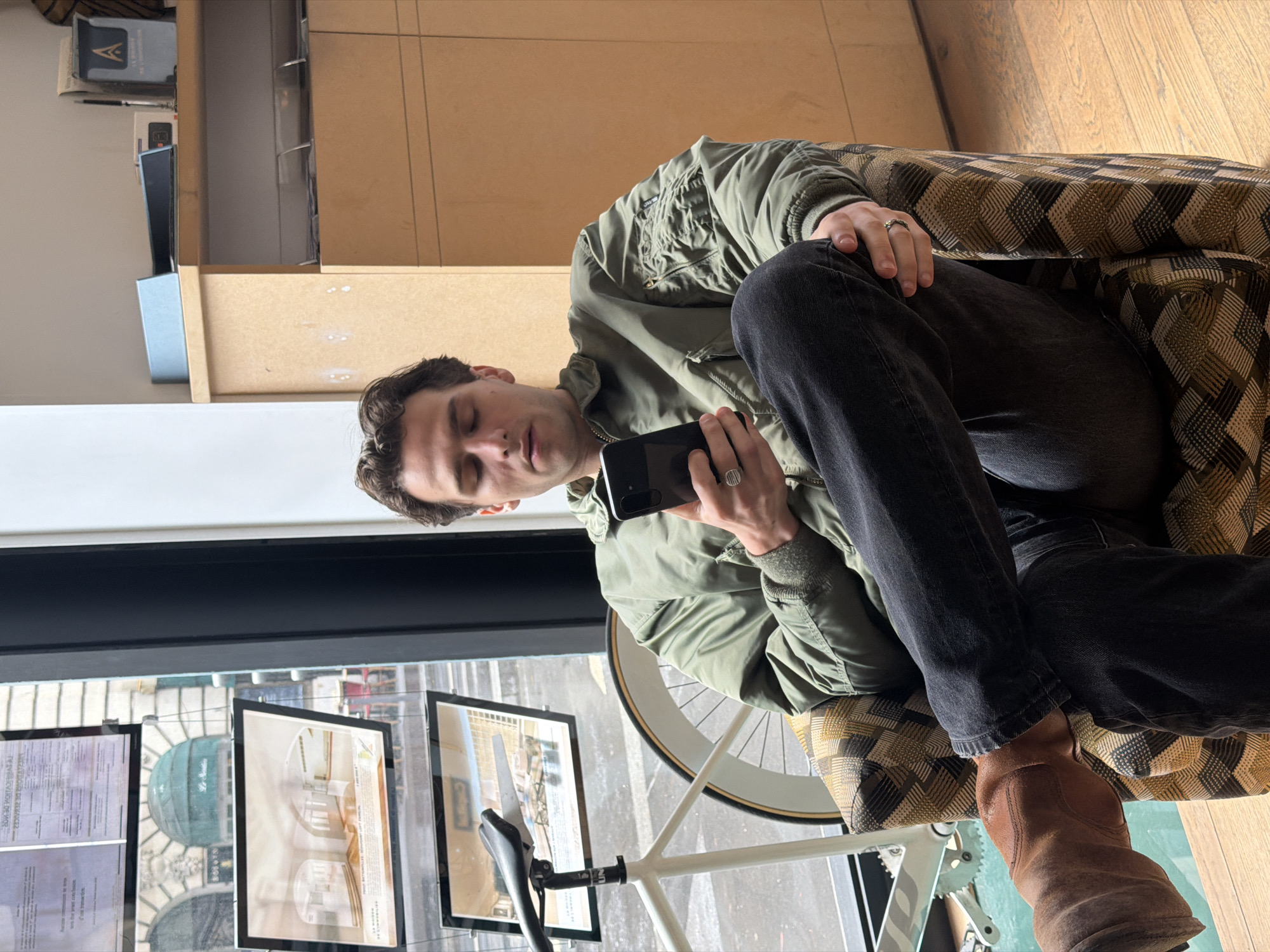 A man is sitting in an armchair, looking at a smartphone, in a building in Paris, France. He is wearing a green jacket, black jeans, and brown boots. The armchair has a patterned fabric. There is a bicycle next to the armchair. The man is wearing two rings. There are real estate advertisements displayed in the window.