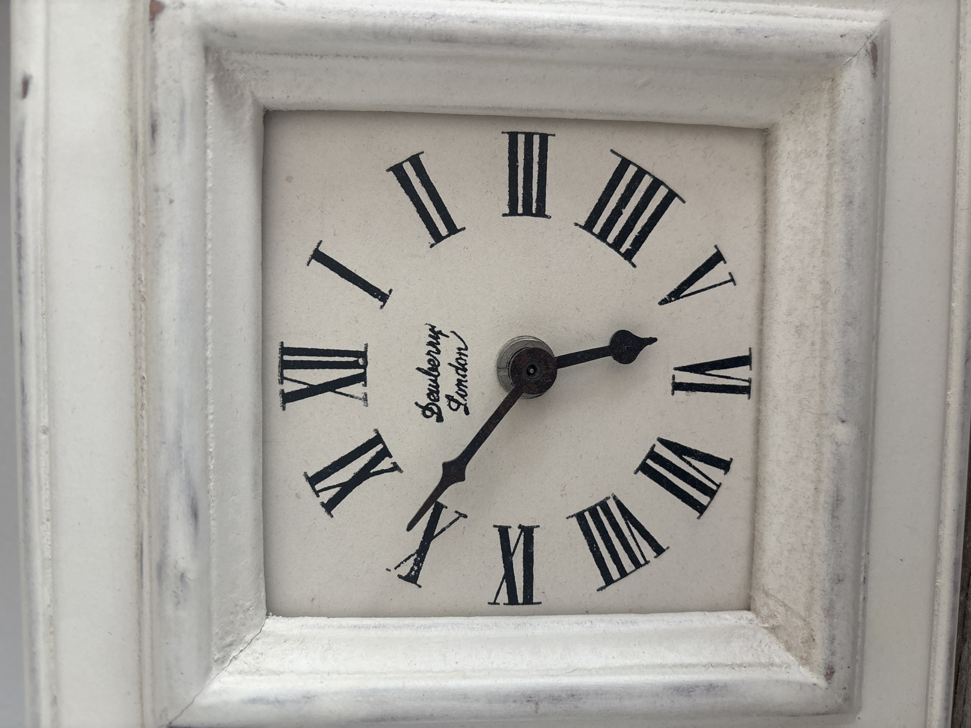 The image shows a close-up of a distressed white wooden framed clock with a cream-colored face. The clock face features black Roman numerals and a brand name "Dewberry London" in script. The hour hand is pointing at approximately 4 o'clock, and the minute hand is pointing at approximately 8 minutes past the hour. The clock appears to be a decorative tabletop or wall clock. The scene is devoid of any identifiable environmental context that would suggest a specific location in Dubai, United Arab Emirates. There are no people, actions, or interactions depicted.