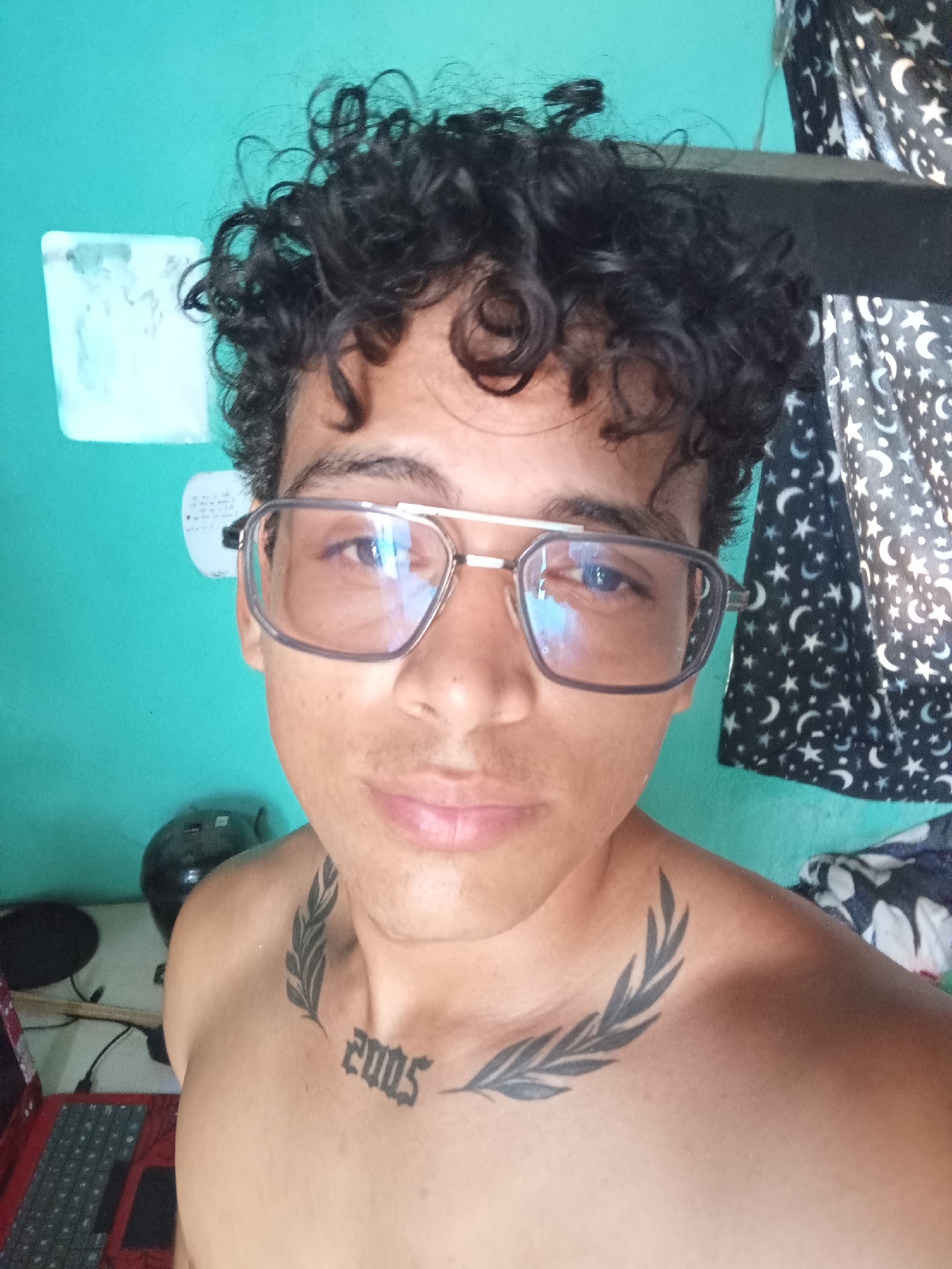 A close-up, self-taken photograph shows a young person with curly black hair and eyeglasses. The person is shirtless, revealing tattoos on their chest and collarbone, including the number "2005" and a laurel branch design. The background features a turquoise wall, a black helmet, a laptop, and a patterned curtain. The image appears to have been taken indoors, possibly in a bedroom, in the city of Altagracia, Venezuela.