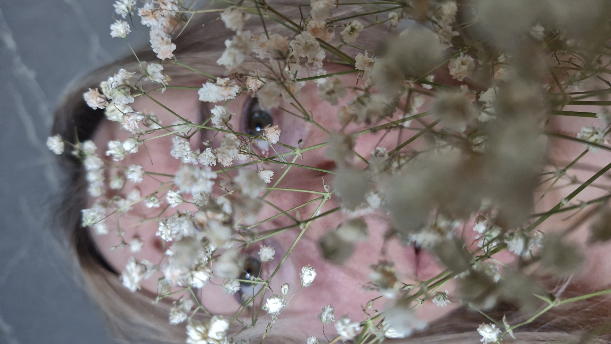 A close-up, slightly rotated, upward-facing shot shows a woman's face partially obscured by a bouquet of small white flowers with thin green stems. Only her eyes, part of her forehead, and cheeks are visible behind the flowers. Her eyes are light brown. She has light blonde or grey hair visible at the sides of the frame. The background appears to be a grey textured surface. The image does not show any recognizable elements to place it in Otchor, Russia or provide any other contextual details.