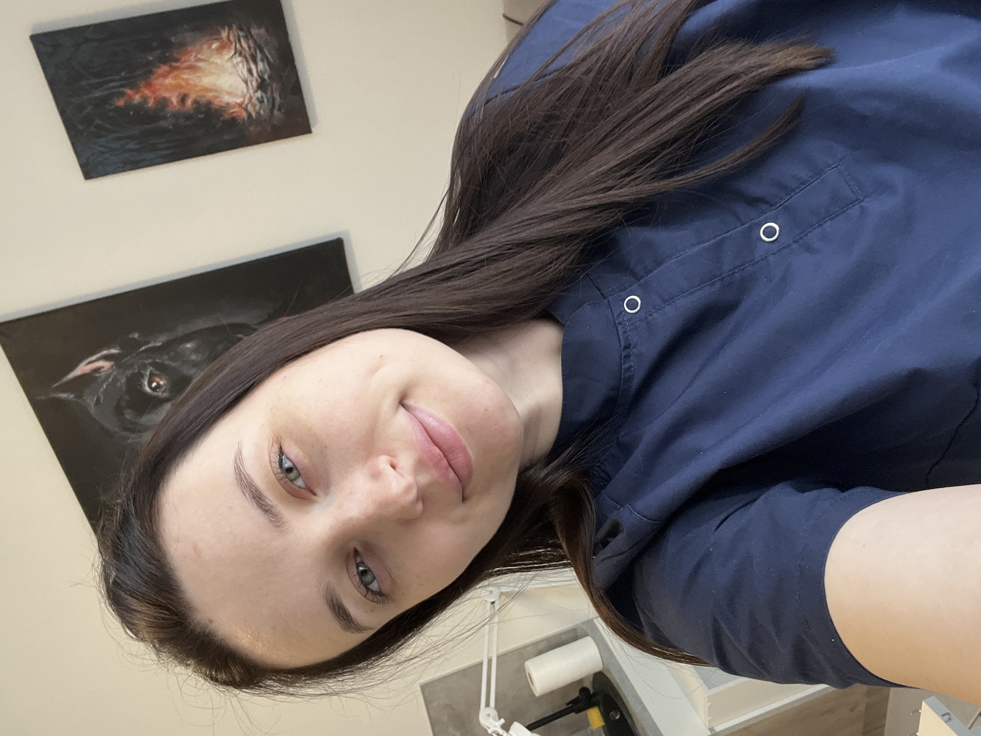 A woman with long, dark hair and blue eyes is wearing a navy blue top with two visible button closures on a pocket. She is smiling and looking directly at the camera. In the background, there are two framed paintings on a light-colored wall. One painting appears to depict fire or flames, while the other seems to show a black animal, possibly a dog or panther. A machine with a roll of white paper can be partially seen in the lower section of the frame. The location is Tyumen, Russia.