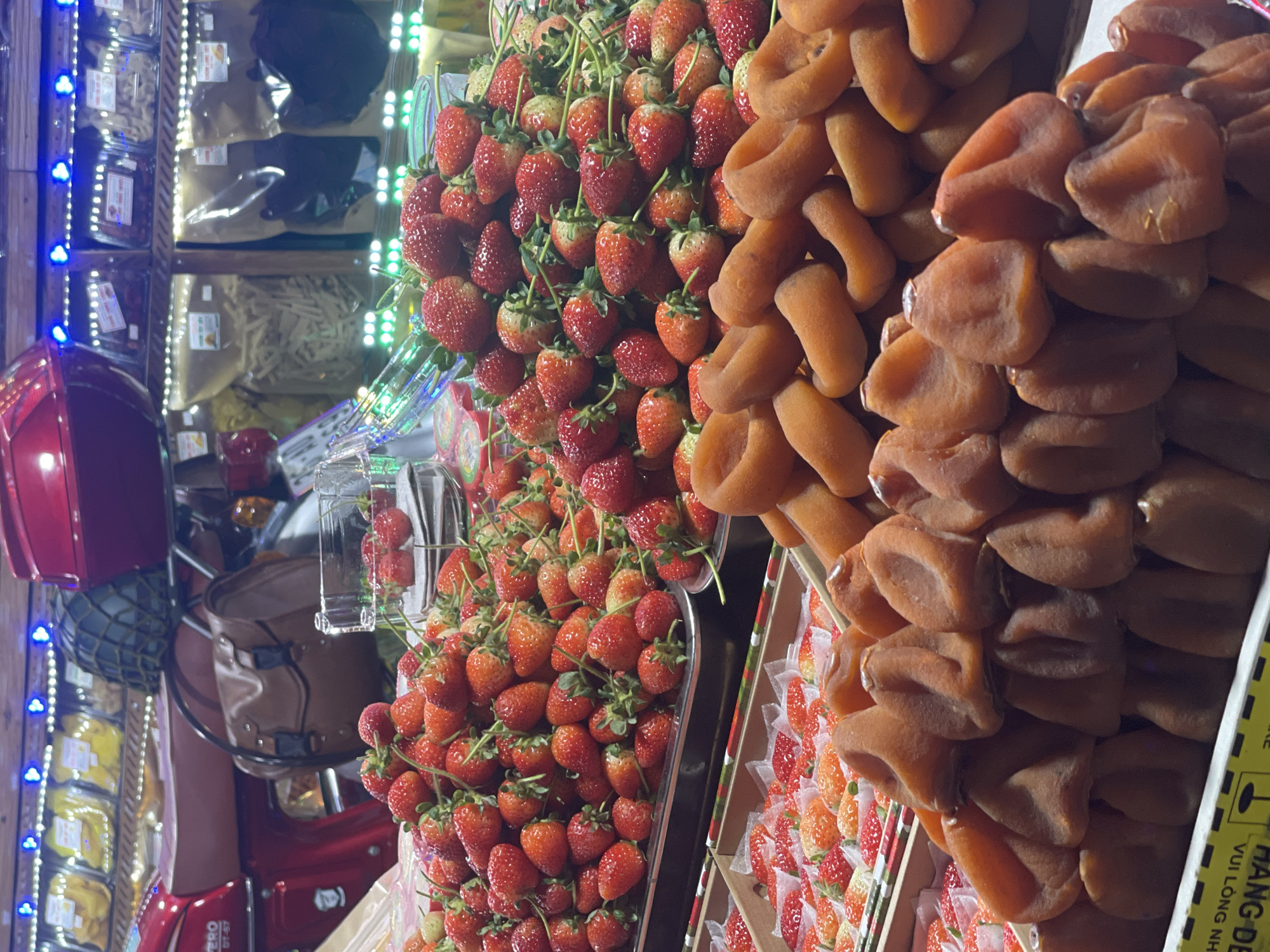 The image captures a vibrant street scene in Da Lat, Vietnam, showcasing a vendor's stall laden with fresh produce. The stall is part of a motorized vehicle, likely a scooter, which also features storage and display cases.
Prominently displayed are piles of fresh strawberries, bright red and green with their stems intact, and dried persimmons. The persimmons are stacked high in organized layers.
The scooter is equipped with a red helmet affixed to its body. The storage and display cases on the scooter contain various packaged foods, visible in the background. Strings of blue LED lights illuminate the stall, enhancing its attractiveness.