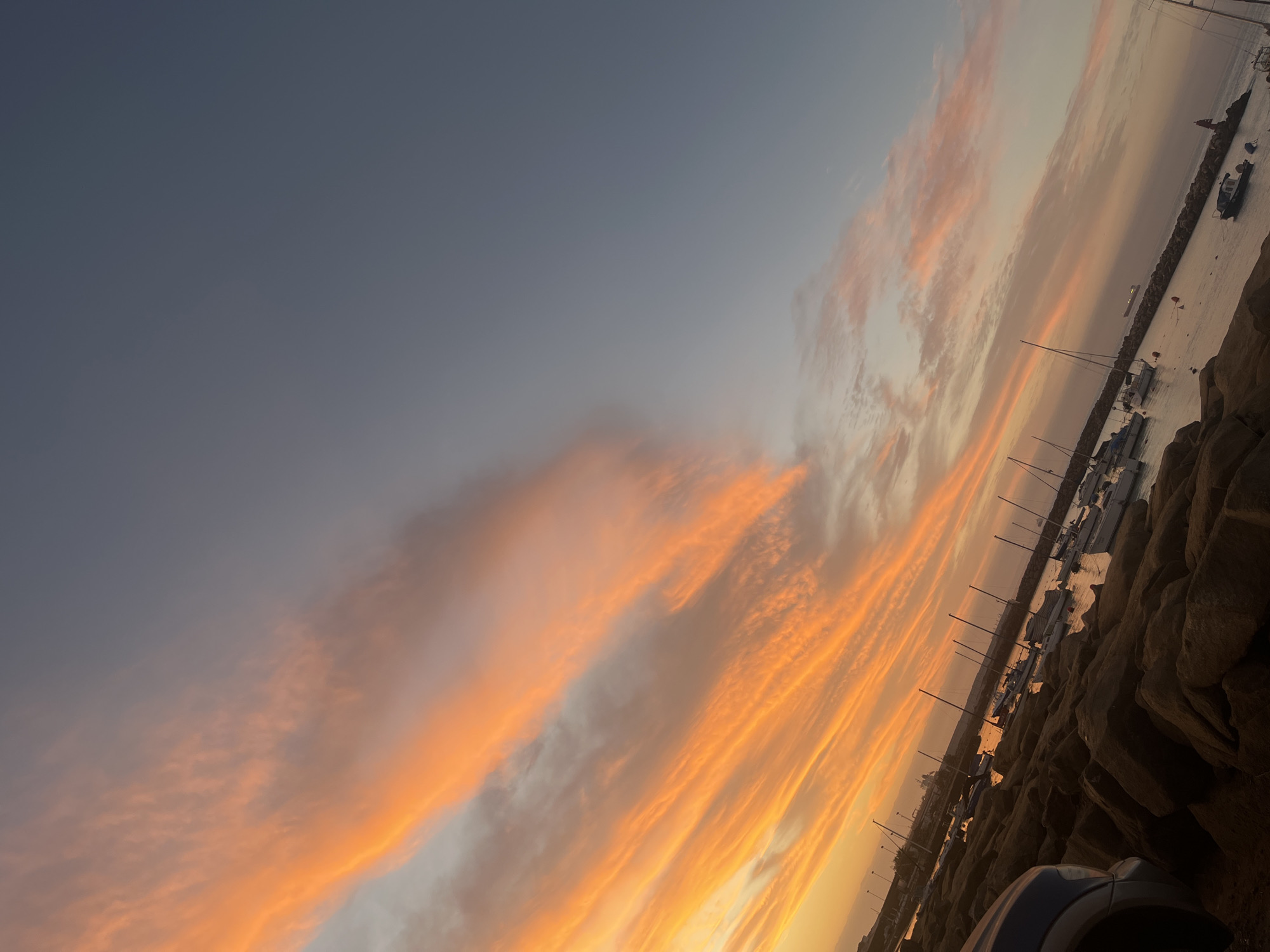 The image shows a dusk scene at a marina in Arica, Chile. The sky is a blend of gray and orange hues due to the sunset. Streaks of orange clouds are prominent, creating a vibrant contrast with the cooler tones in the upper part of the sky.
The marina area features boats, some with visible masts, docked alongside a rocky shoreline or breakwater. The rocks appear dark and coarse, providing a solid boundary between land and water. The water surface reflects the colors of the sky, adding to the atmospheric effect. A portion of a car is visible in the foreground, suggesting the photo was taken from or near a vehicle.