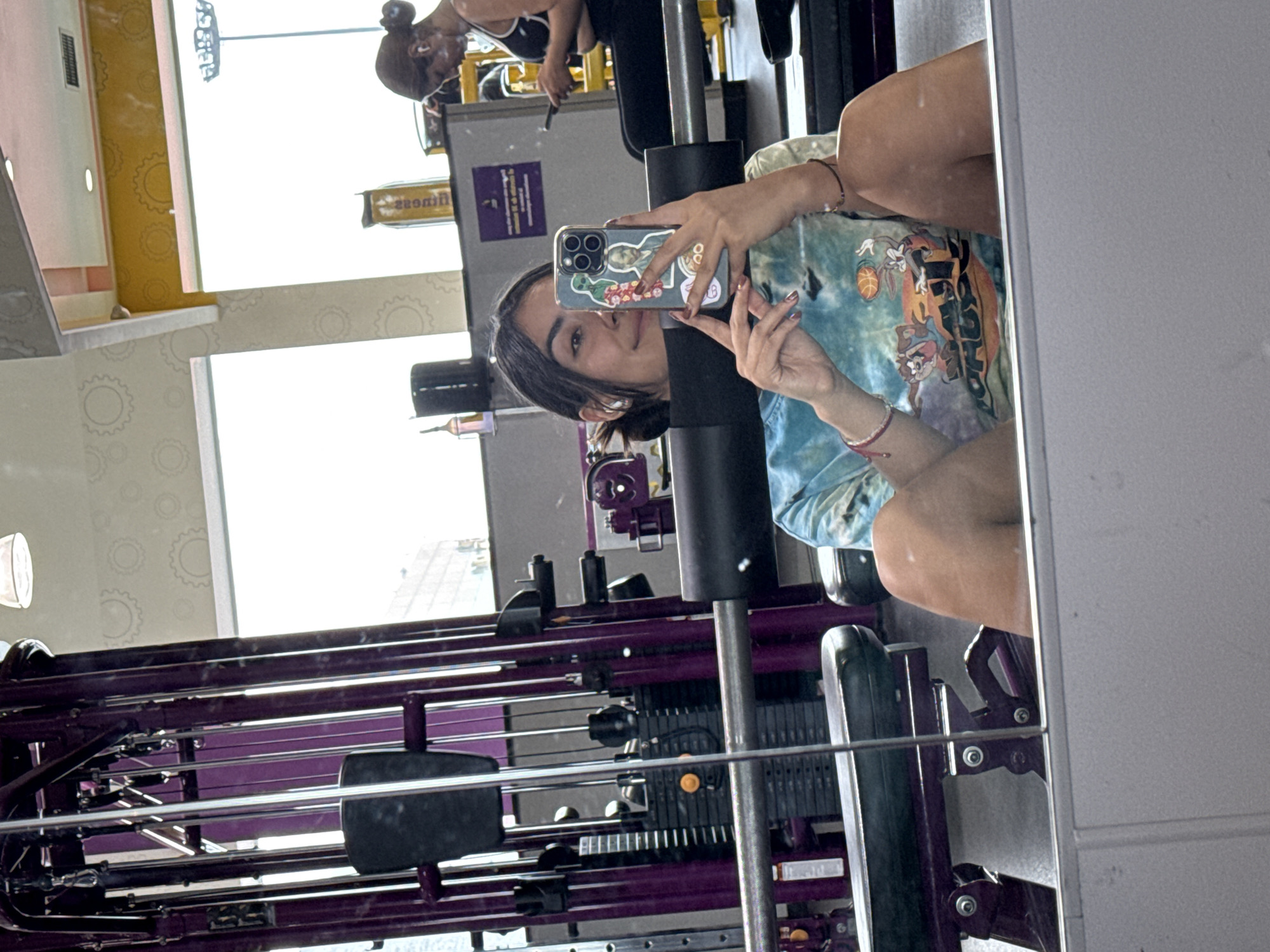 A young woman takes a selfie in a mirror at what appears to be a gym in Puebla, Mexico. The gym features purple exercise equipment. The woman has brown hair, light skin, and is wearing a t-shirt. She is holding a cell phone with a decorated case. Another person is indistinctly visible in the background, working on exercise equipment. The gym's interior features yellow and white walls. Outside the window, a cityscape is visible.