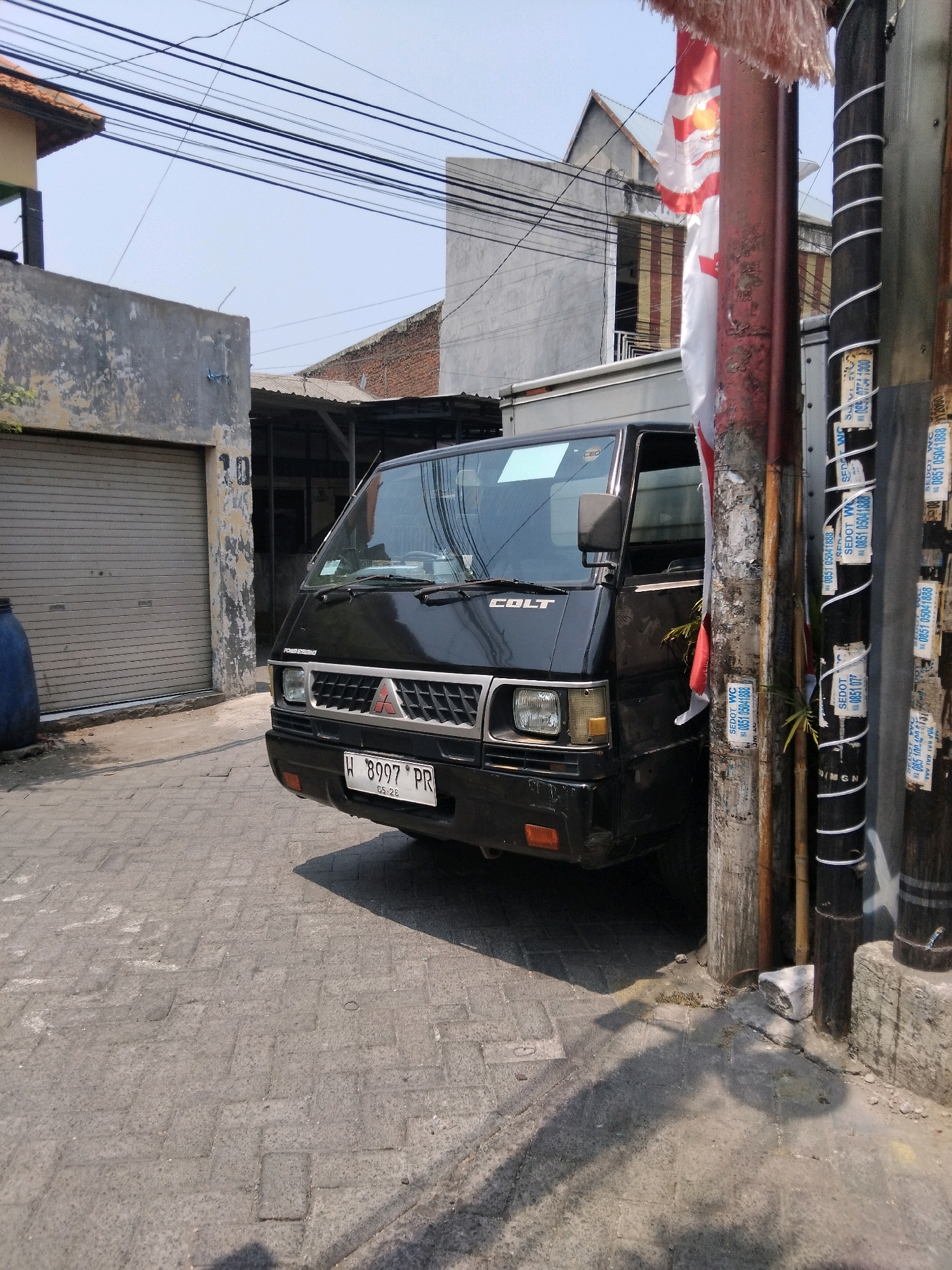 An outdoor, eye-level, medium shot captures a black Mitsubishi Colt light truck parked on a paved street in Kepuh Kiriman, Indonesia. The truck faces slightly left, with its front grille and headlights prominently visible. The license plate reads "W 8997 PR". To the left of the truck is a gray roll-up garage door. To the right, utility poles are adorned with various stickers and notices. A red and white Indonesian flag hangs on a pole, partially obscuring the view of a building behind it. The sky is clear and blue, indicating daytime. The overall scene depicts a typical urban streetscape in Indonesia.