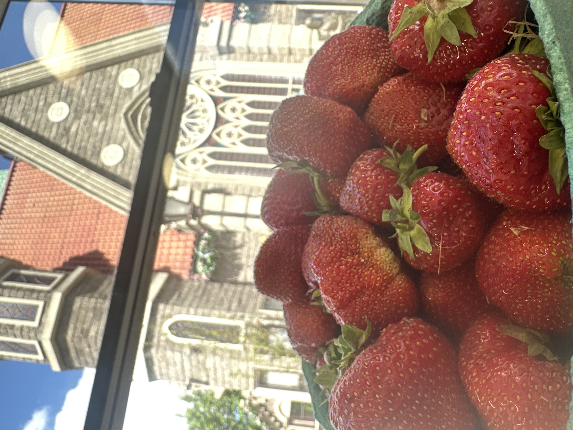 This image depicts a close-up view of a basket filled with fresh, ripe strawberries. The strawberries are a vibrant red, with visible seeds and green leafy tops. They are packed tightly together, overflowing from a green cardboard container. In the background, out of focus, is a building with a distinctive architectural style, featuring a red-tiled roof, stone facade, and arched windows. The sky above is a bright blue with some clouds. The overall impression is one of a fresh, natural product set against a charming, possibly historic, backdrop. The image does not provide any specific information to suggest it is located in Moscow, Russia.