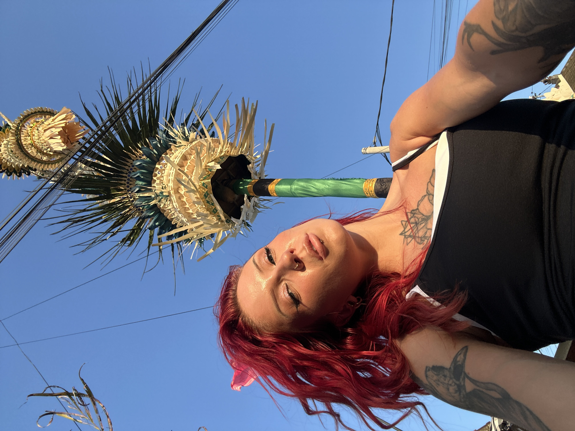 A young woman with bright red hair stands outdoors against a bright blue sky. She is looking up and to the left, with her head tilted back and her lips slightly parted. She is wearing a black tank top with white trim and has tattoos on her arms and chest. Her hair is styled in loose waves and falls over her shoulders.
In the background, a tall, green pole is visible, adorned with decorative elements made of straw and leaves. This appears to be a Balinese ceremonial umbrella, commonly seen during religious festivals. Above the umbrella, numerous black wires crisscross the sky, indicating the presence of utility infrastructure, likely in an urban or semi-urban setting. The sunlight casts a warm glow on the woman's face and arms. The overall scene suggests a bright, sunny day in North Kuta, Indonesia, possibly during a local celebration or event.