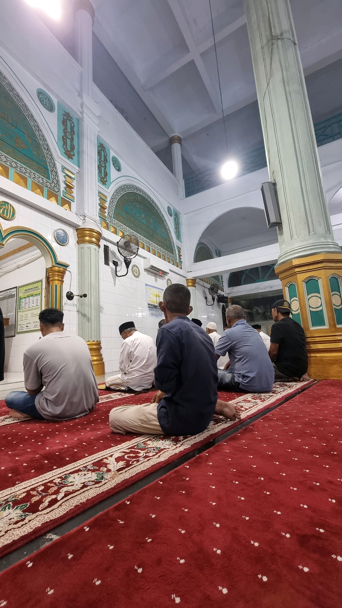 A group of men are praying in a mosque in Puloara, Indonesia. They are seated on a red carpet with a floral pattern. The mosque has white tiled walls with decorative green and gold accents. Large white pillars with gold trim support the ceiling. A fan is mounted on the wall.