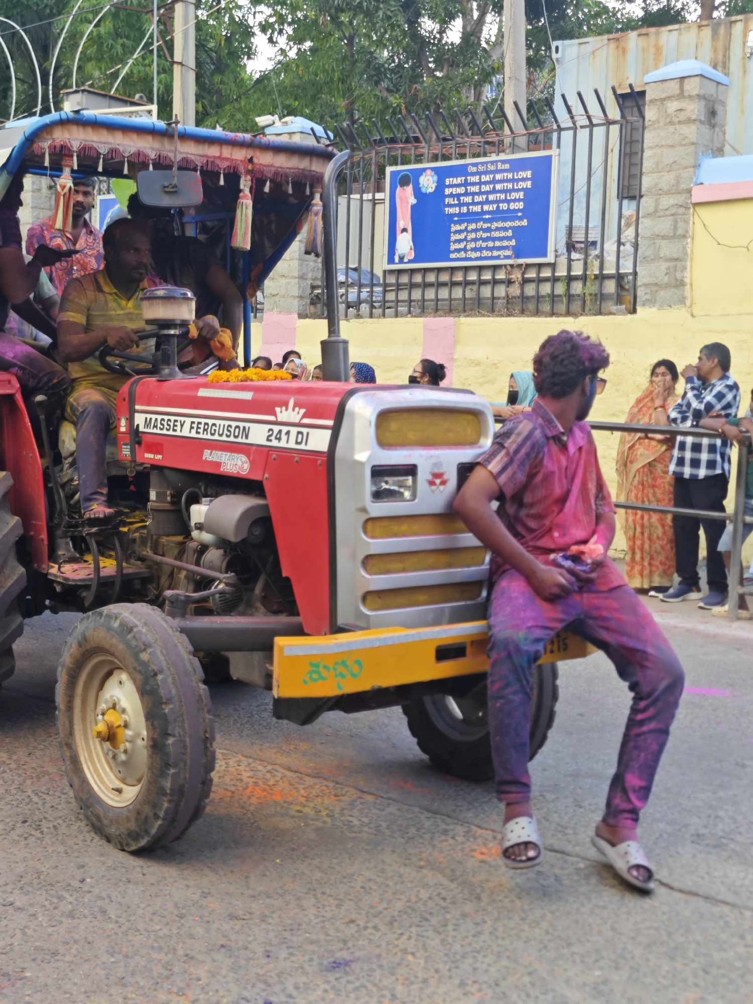 In Puttaparthi, India, a red Massey Ferguson 241 DI tractor is depicted, adorned with colorful tassels and flower garlands on its canopy and hood, suggesting a festive occasion, possibly a procession or celebration like Holi, given the presence of colored powder on people and the environment. A man, partially covered in vibrant colored powder, sits on the side of the tractor's yellow-painted front loader, facing away from the camera. He is wearing a patterned shirt and pants, with some of the powder adhering to his face and hair. The tractor is stationary on a paved road, with other individuals visible in the background. Some are watching the scene, while others appear to be involved in the festivities. Behind the tractor, a white metal fence encloses an area with a blue sign containing English and Telugu text, alongside a graphic of a robed figure. The general atmosphere suggests a lively, communal gathering.